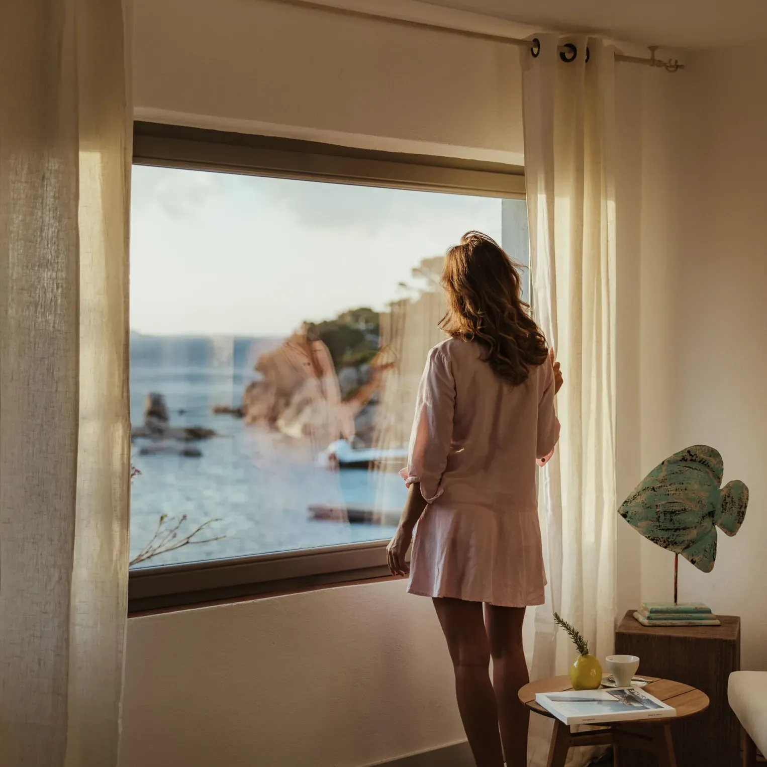Une femme en robe regarde par la fenêtre une vue sereine sur l’océan avec des falaises rocheuses. Une lumière douce remplit la pièce, créant une atmosphère paisible et contemplative.