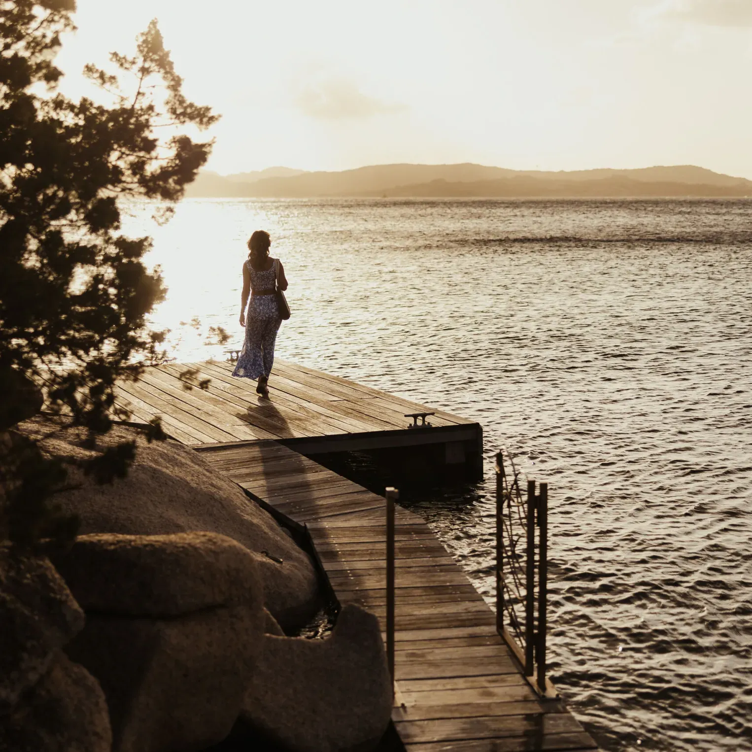 Une personne en robe bleue marche le long d’un ponton en bois vers l’eau. Le soleil se couche à l’arrière-plan, projetant une lumière chaude sur une scène paisible.