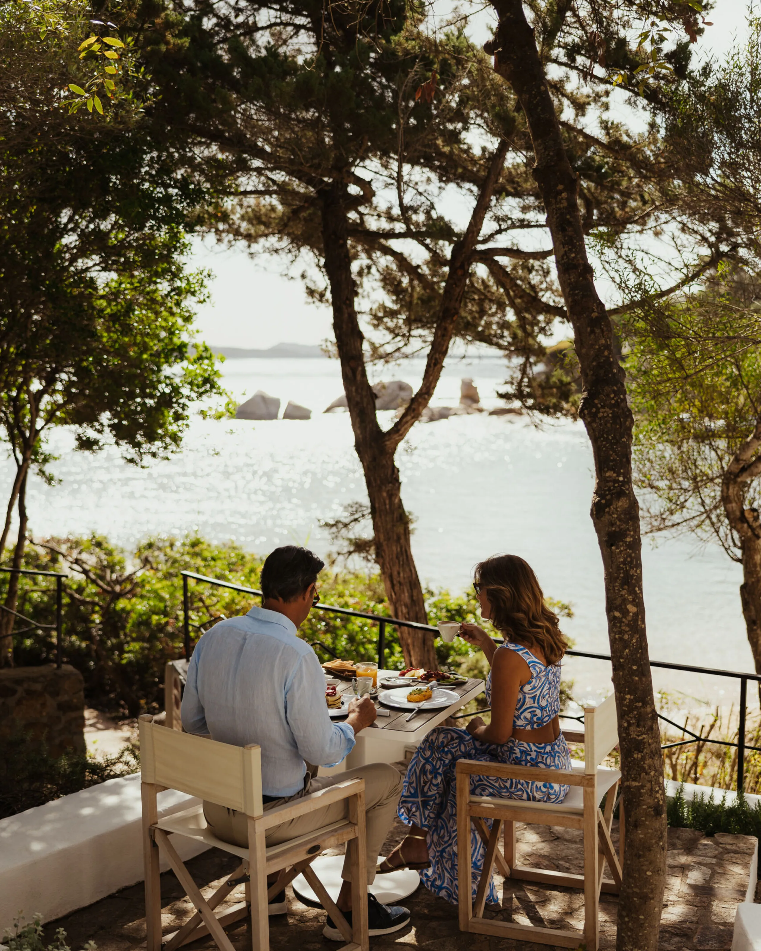 Un couple prend son petit-déjeuner en plein air sous les arbres, avec vue sur un paysage côtier paisible. La lumière du soleil scintille sur l’eau, créant une atmosphère calme et romantique.