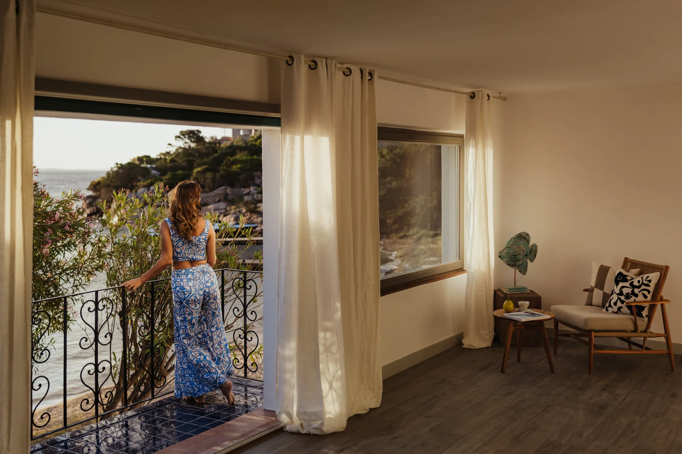 Woman in a blue dress stands on a balcony overlooking the ocean at sunset, casting a serene and contemplative mood. Room features a cozy chair and table.