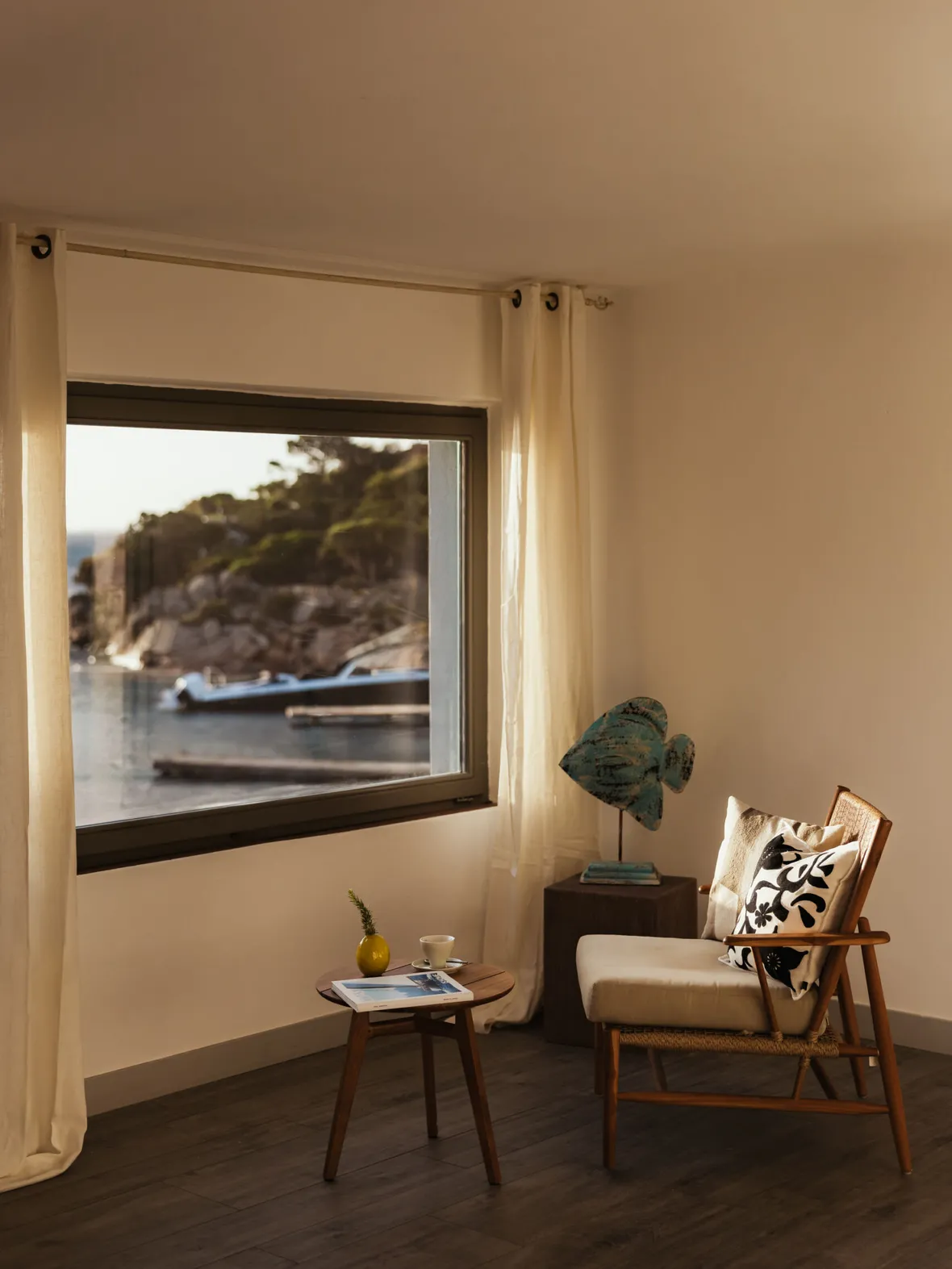Cozy room with a mid-century chair and side table near a large window revealing a serene seaside view. Soft lighting and neutral tones create a calming ambiance.