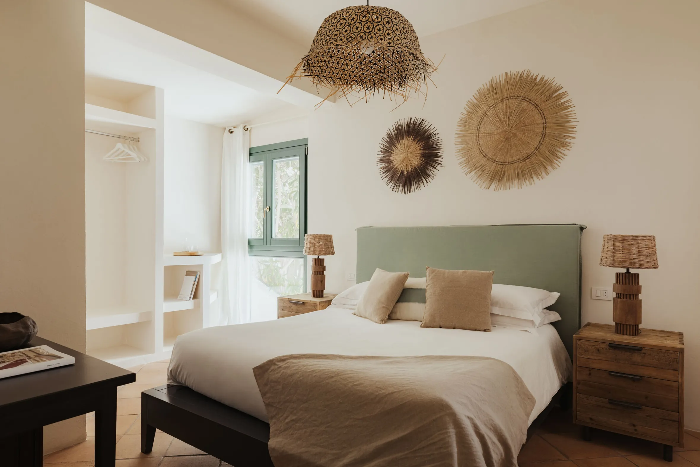 Minimalist bedroom with a cozy bed and green headboard, flanked by wooden nightstands. Woven wall decor and a rattan light fixture add warmth.