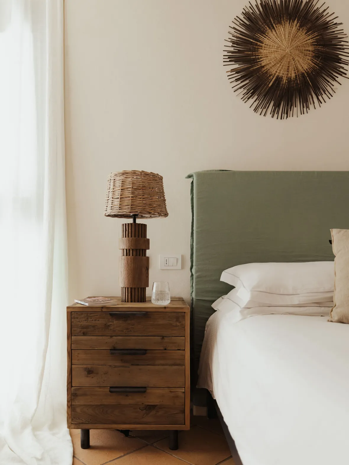 A cozy bedroom scene featuring a wooden nightstand with a wicker lamp, beside a bed with an olive-green headboard. A sunburst wall decor adds charm.