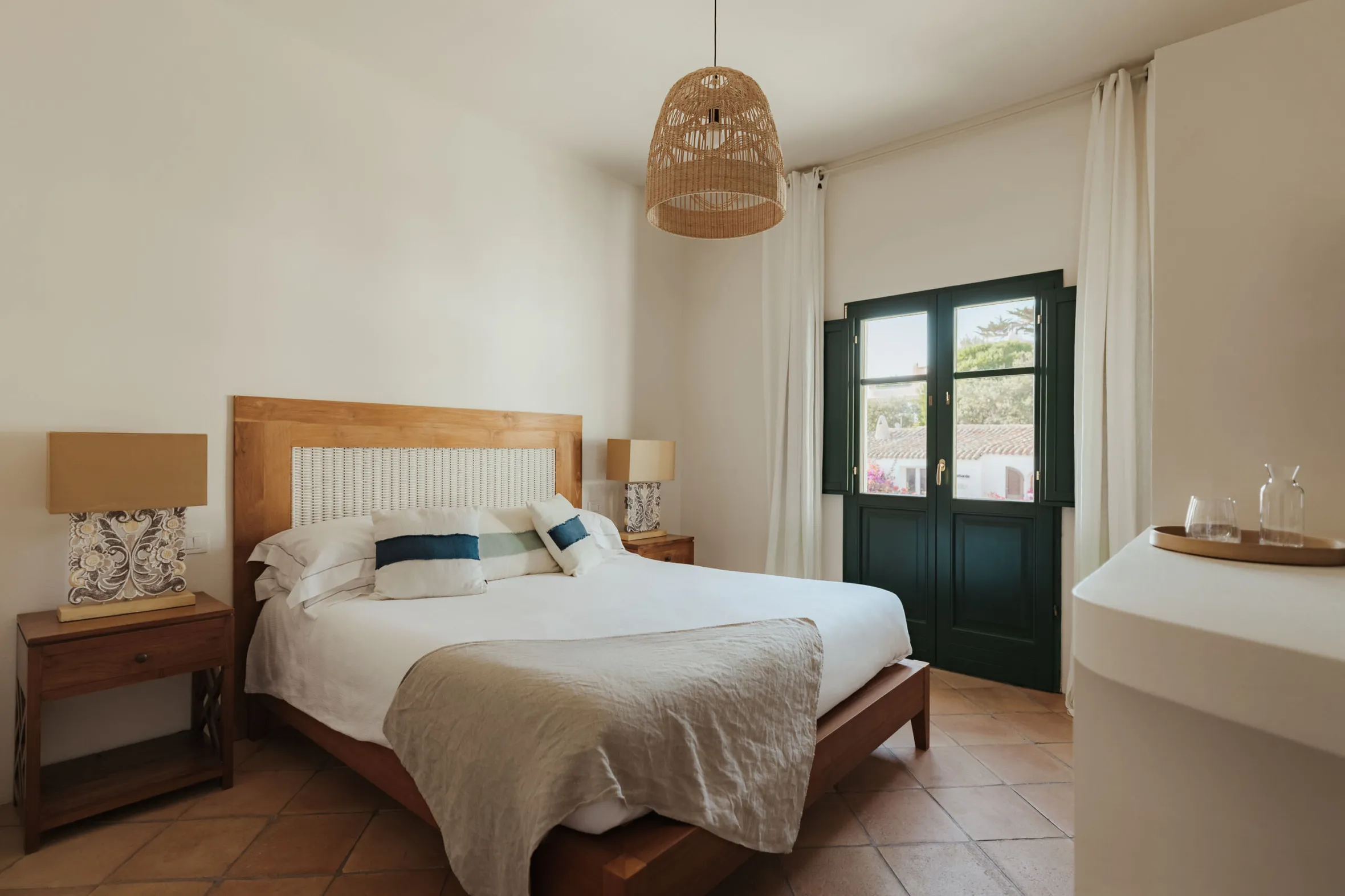 Cozy bedroom with a wooden bed, white and blue pillows, wicker lamp, and rustic nightstands. Sunlight streams through a window with green shutters.