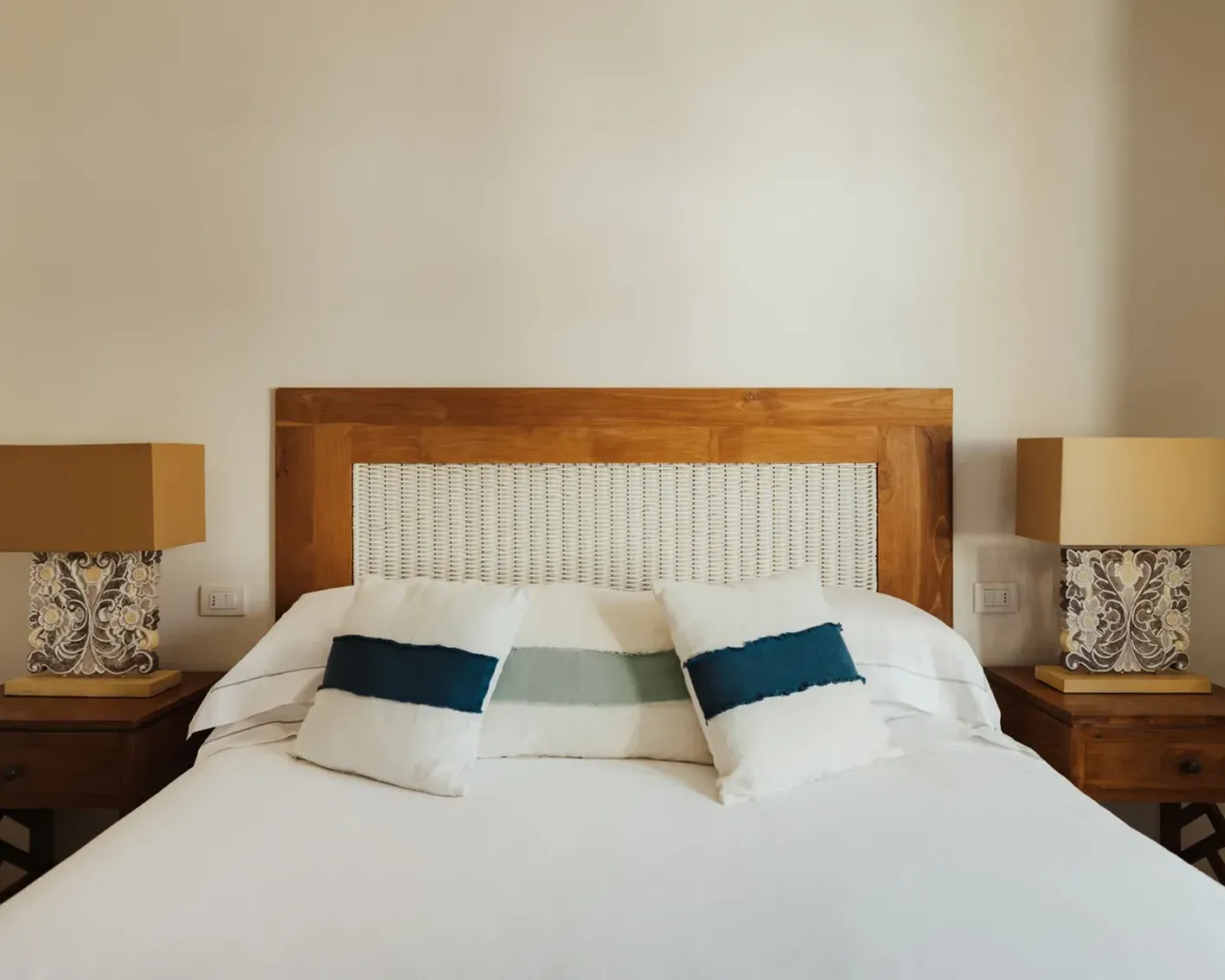 A cozy bedroom features a bed with a woven wooden headboard, three accent pillows in blue and white, and two nightstands with patterned lamps, creating a calm ambiance.