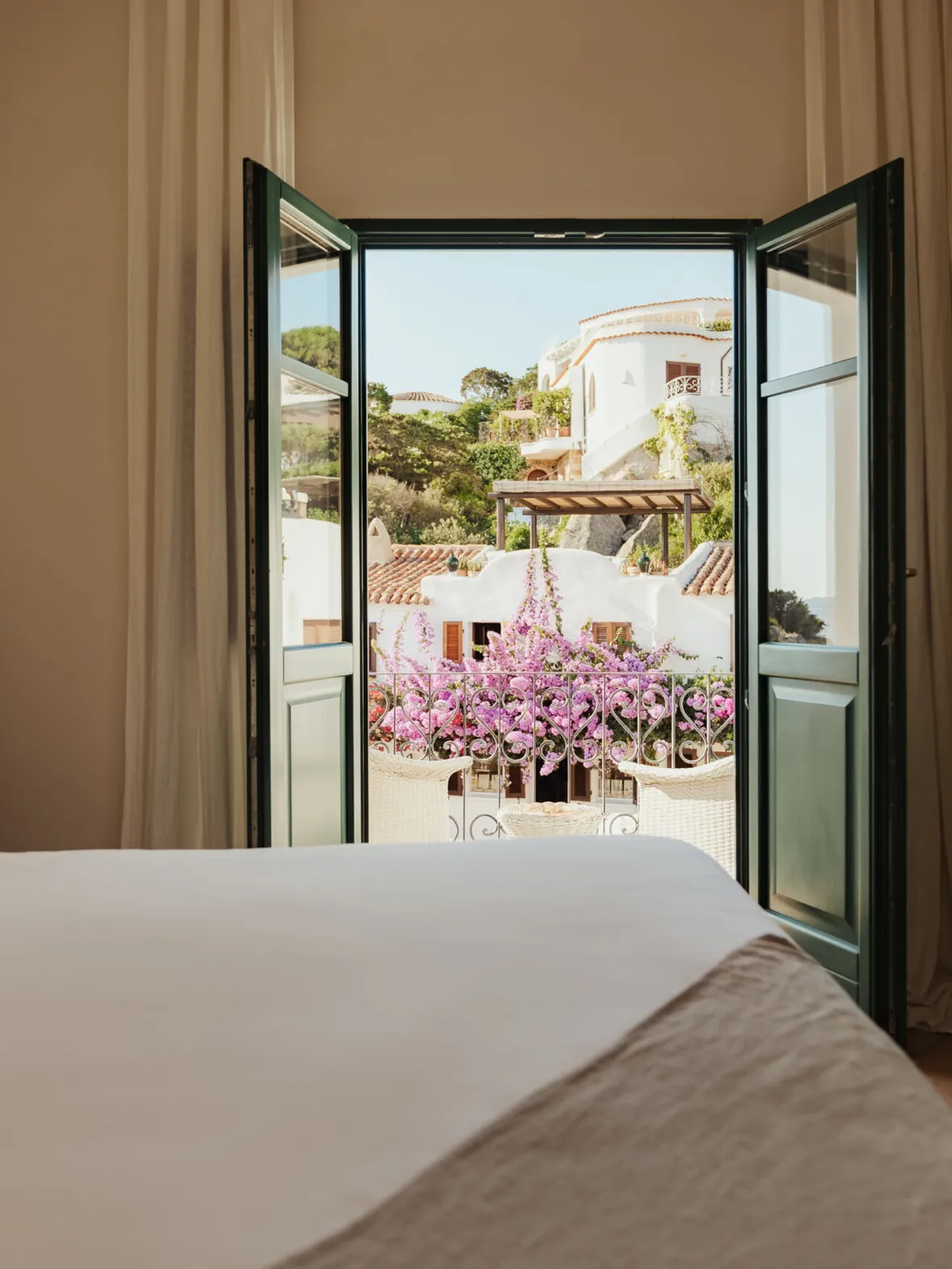Bright, airy bedroom with an open door leading to a balcony filled with vibrant pink flowers. View of charming Mediterranean architecture outside.
