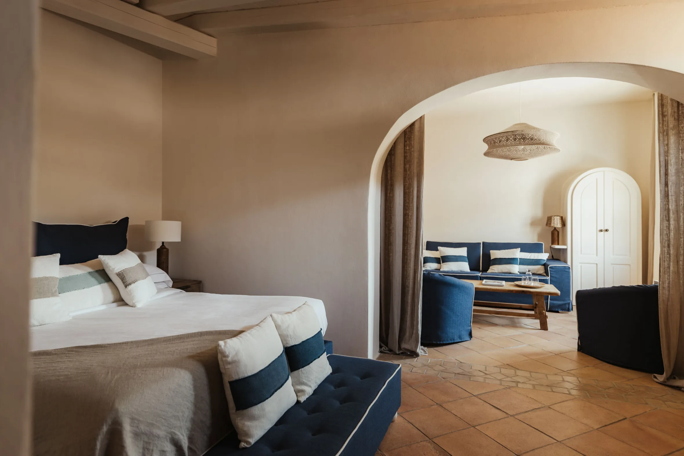 Spacious bedroom with neutral tones, featuring a bed with striped cushions, cozy lounge area with blue sofas, and arched doorway; calm and inviting.