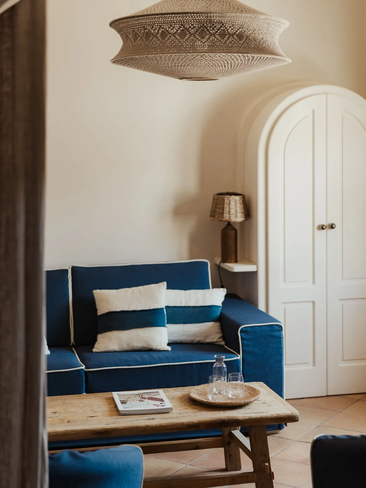 Cozy living room with a blue sofa, striped cushions, and a rustic wooden coffee table. A woven pendant light hangs above, enhancing the calm ambiance.