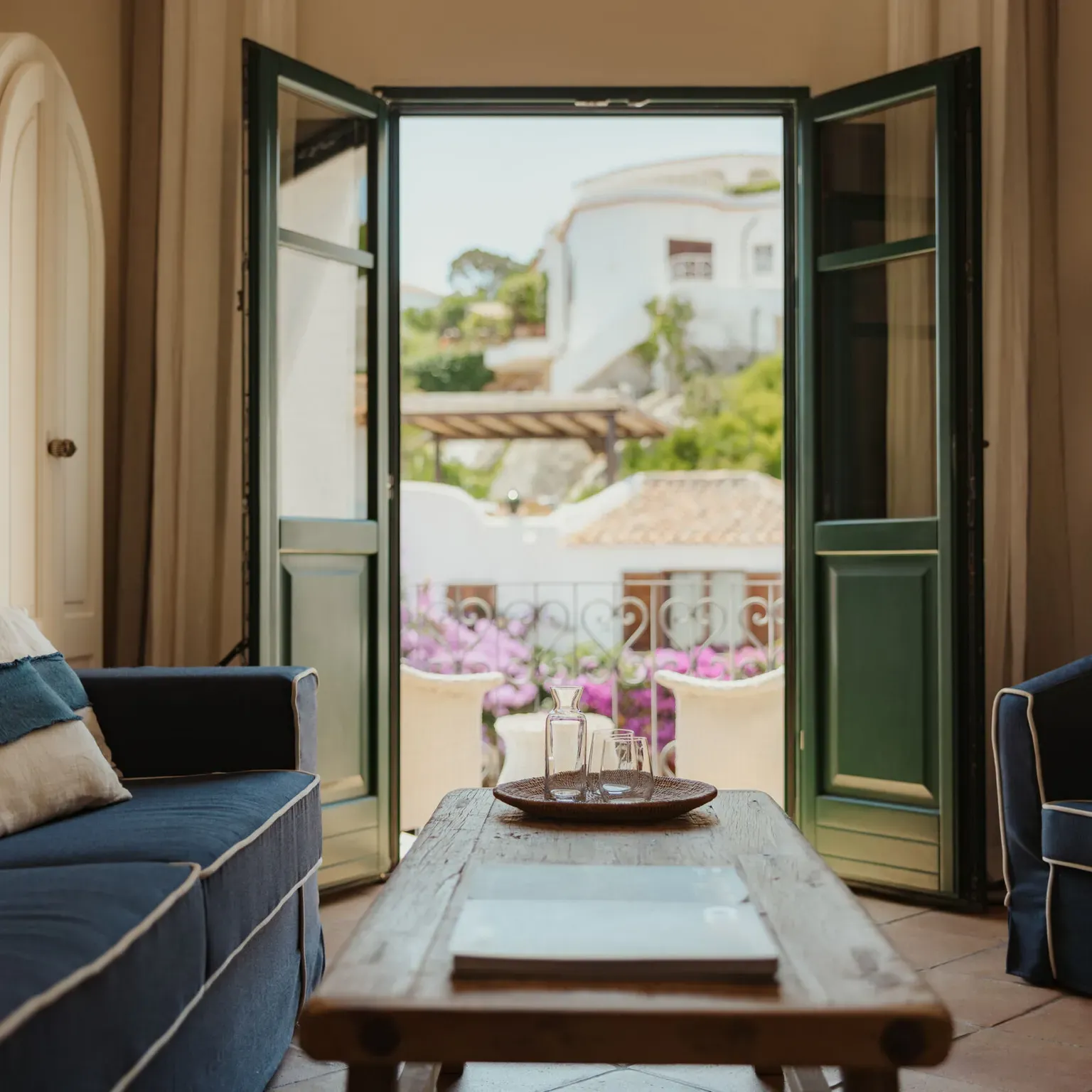 Cozy living room with blue sofas, wooden table, and open French doors leading to a sunny balcony with pink flowers and white Mediterranean-style buildings. Airy and inviting atmosphere.