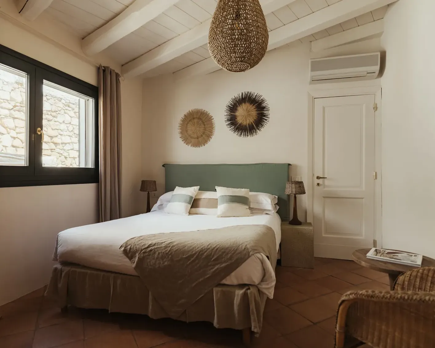 Cozy bedroom with a double bed, green headboard, and striped pillows. Rustic decor includes woven art on a cream wall, wicker furniture, and soft lighting.