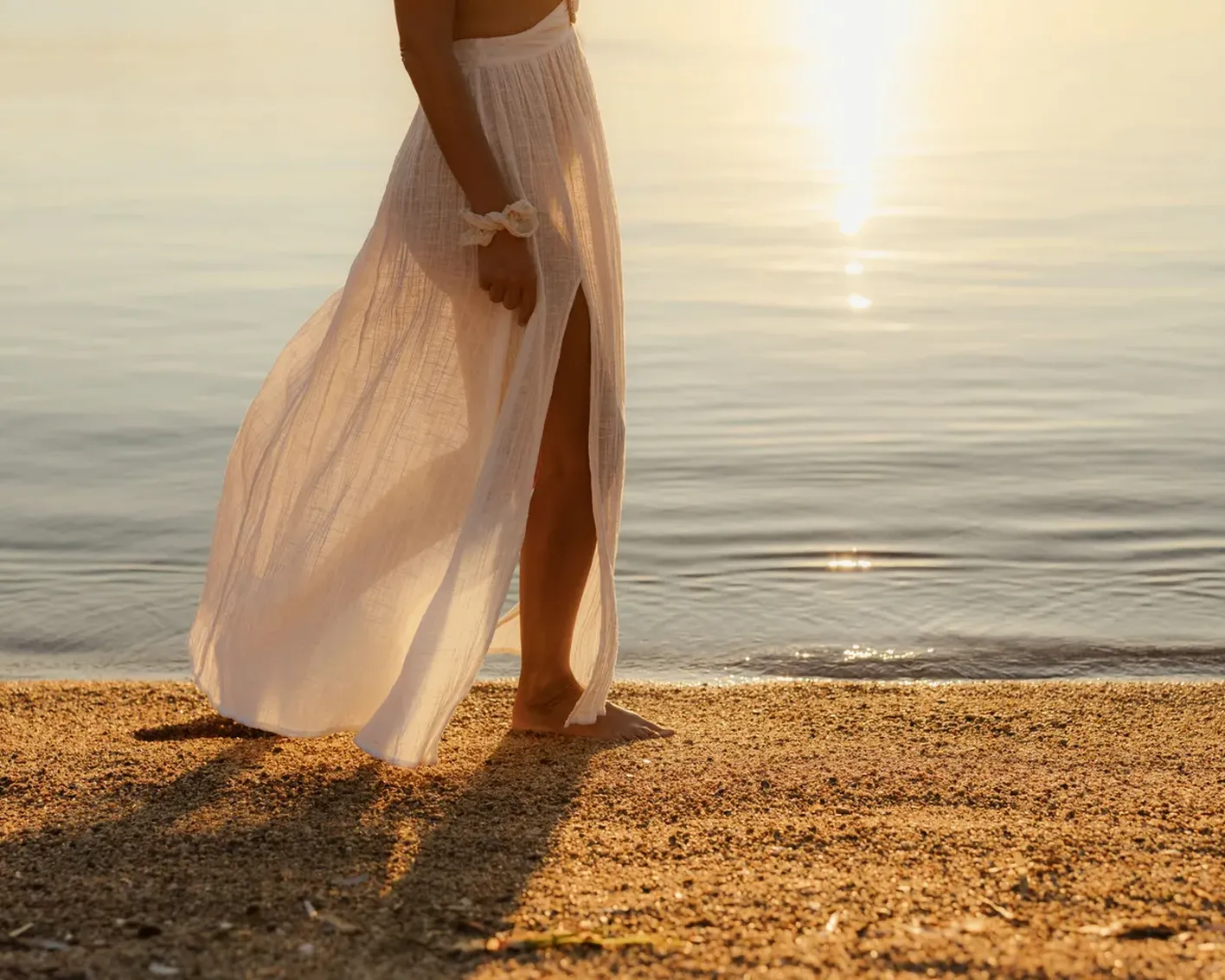 A woman in a flowing white skirt walks barefoot on a sunlit beach, with gentle waves reflecting the golden sunset, creating a serene and peaceful mood.