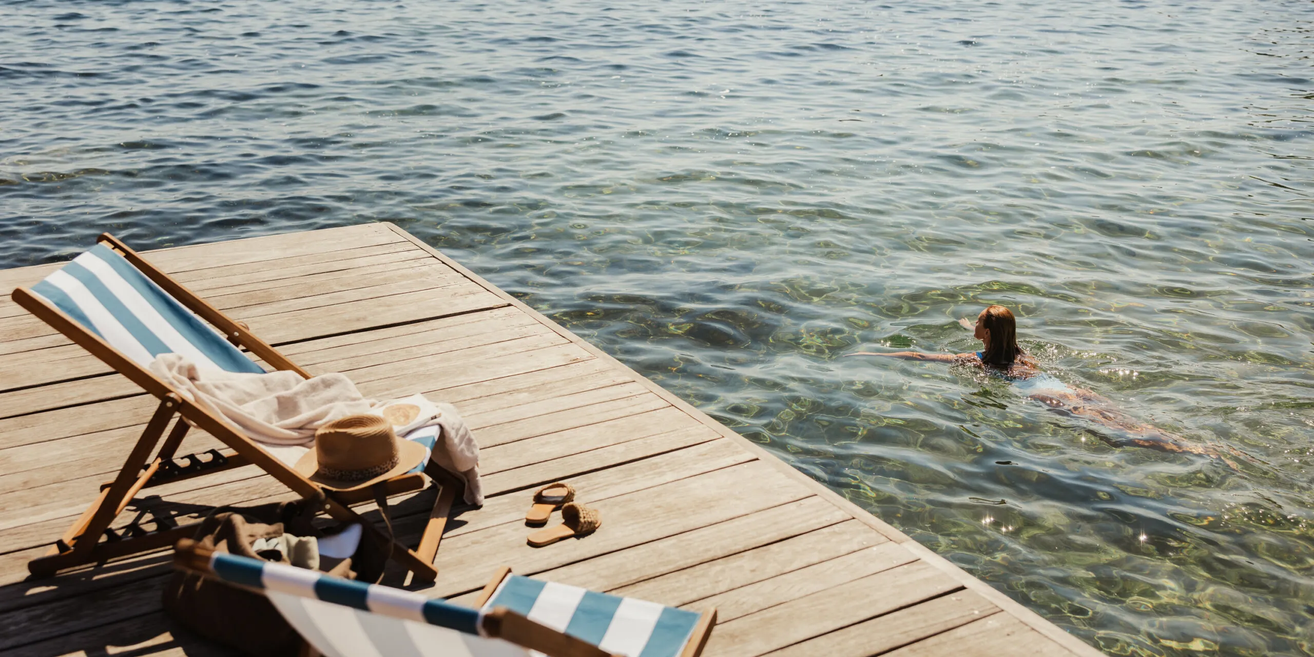 Two striped deck chairs on a wooden dock overlooking clear blue water, with a person swimming nearby. A relaxed, sunny beach vibe is conveyed.
