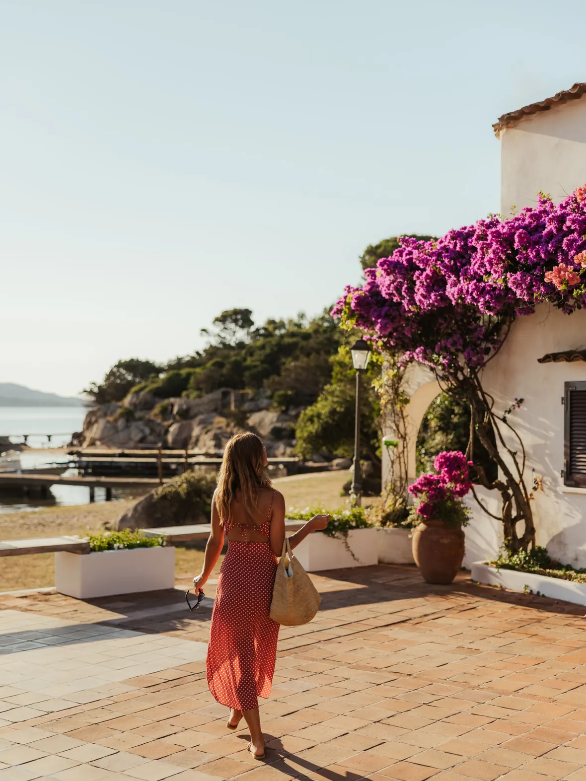 A woman in a red sundress walks by a seaside villa adorned with vibrant purple bougainvillea. The scene is sunny and tranquil, evoking a relaxed, summery vibe.