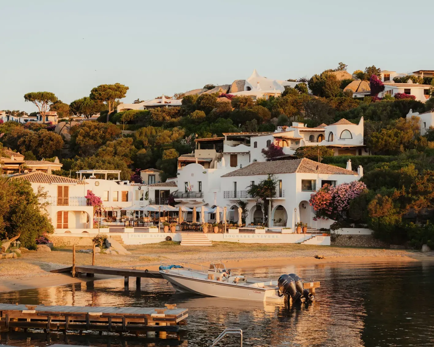 Coastal village at sunset with white Mediterranean-style houses on a hillside, surrounded by lush greenery. Boats and docks reflect on calm water.