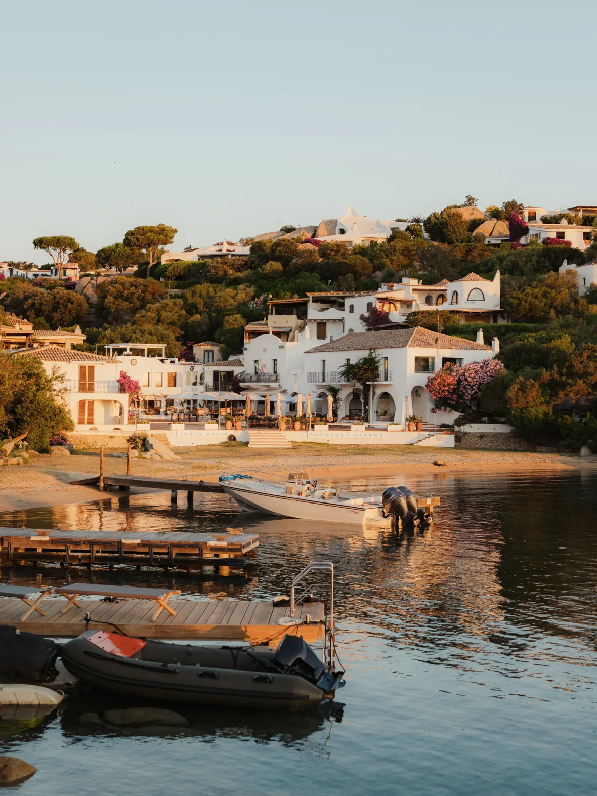 Coastal village at sunset with white Mediterranean-style houses on a hillside, surrounded by lush greenery. Boats and docks reflect on calm water.