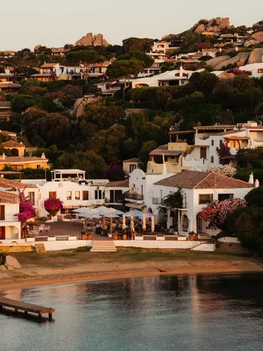 Coastal village at sunset with white villas, terracotta roofs, and vibrant bougainvillea on a hillside. A serene bay and dock are in the foreground.