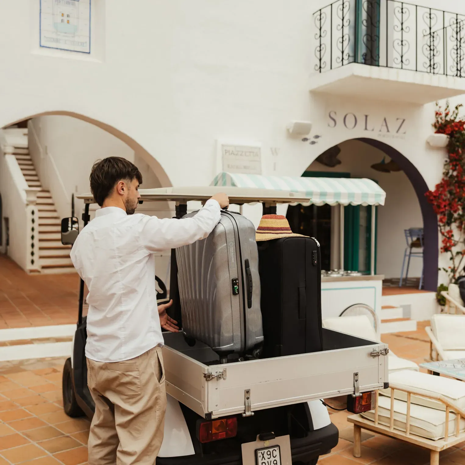 Un homme portant une chemise blanche et un pantalon kaki charge des bagages dans un petit véhicule devant un hôtel de style méditerranéen nommé Solaz. La scène est détendue et ensoleillée.