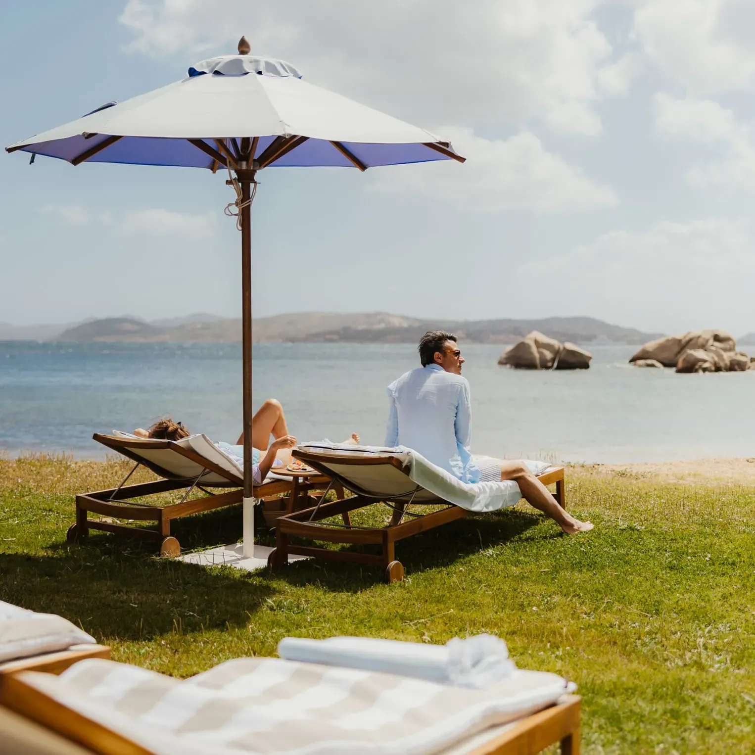 Un couple se détend sur des chaises longues sous un parasol de plage, avec vue sur une mer calme et des îles rocheuses au loin. La scène est paisible et baignée de soleil.