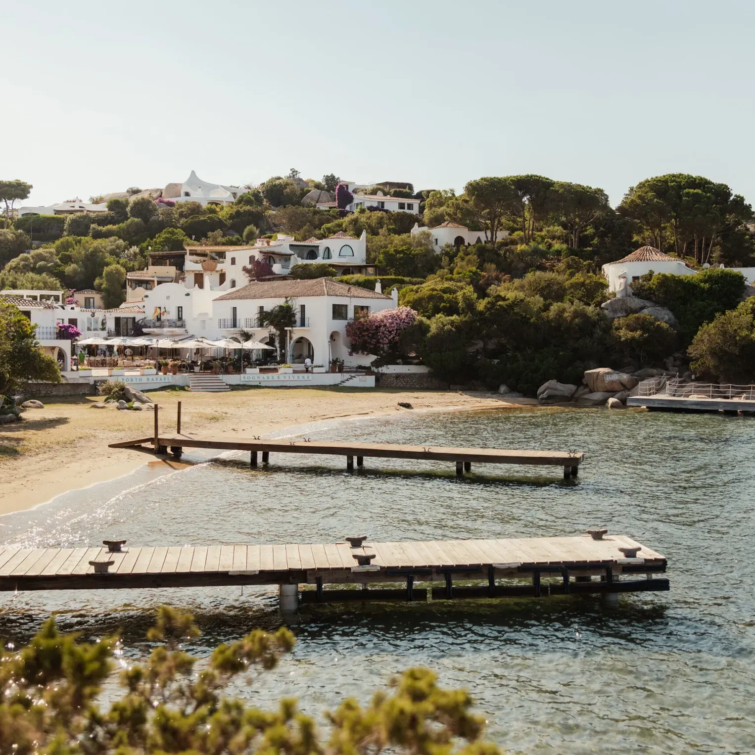 A serene coastal village with white buildings nestled on a green hill, sandy beach, and wooden piers extending over calm blue water under a clear sky.