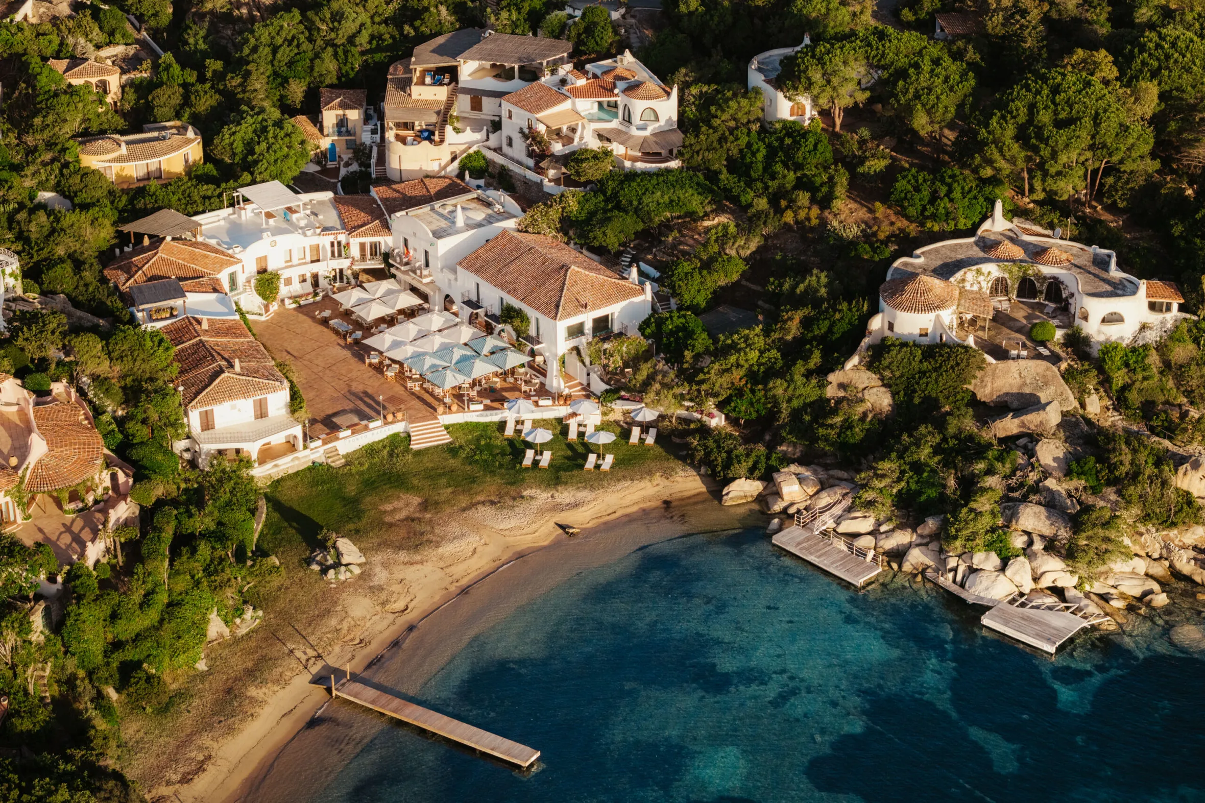 Aerial view of Porto Rafael with whitewashed villas and terracotta roofs nestled among lush greenery. A sandy beach meets crystal-clear blue waters.