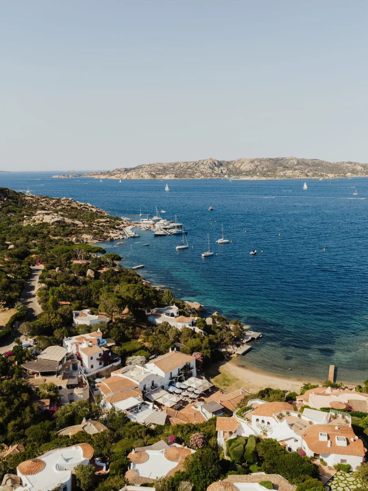 Aerial view of a coastal village with white-roofed houses surrounded by lush greenery. Yachts and sailboats float on the tranquil blue sea. Clear sky.