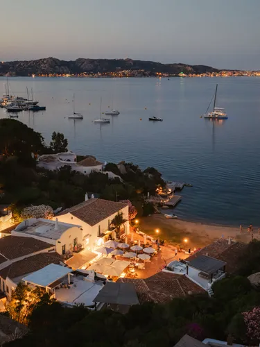 Scena serena di una sera costiera con ville illuminate vicino a una baia tranquilla, dove barche sono ancorate al largo. Luci calde e soffuse creano un’atmosfera pacifica.