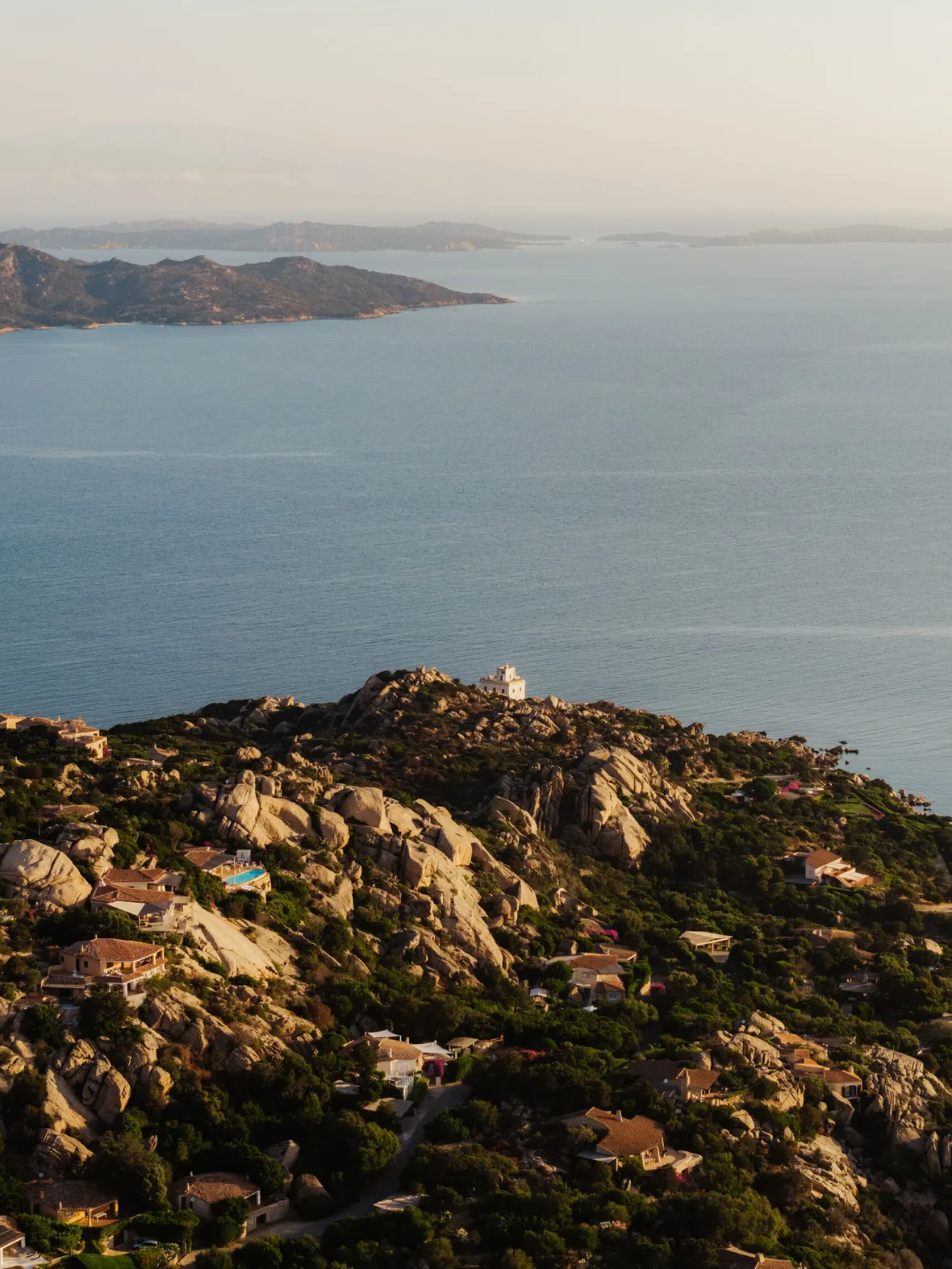 Aerial view of a coastal landscape at sunset, featuring rocky hills dotted with houses. A calm sea and distant islands under a soft, warm light complete the serene scene.
