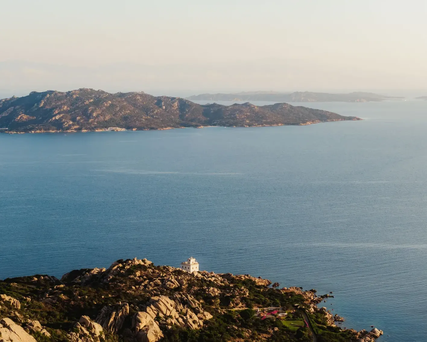 A serene coastal landscape with a rocky foreground and a small white lighthouse, overlooking a calm blue sea. Distant mountains provide a tranquil backdrop.
