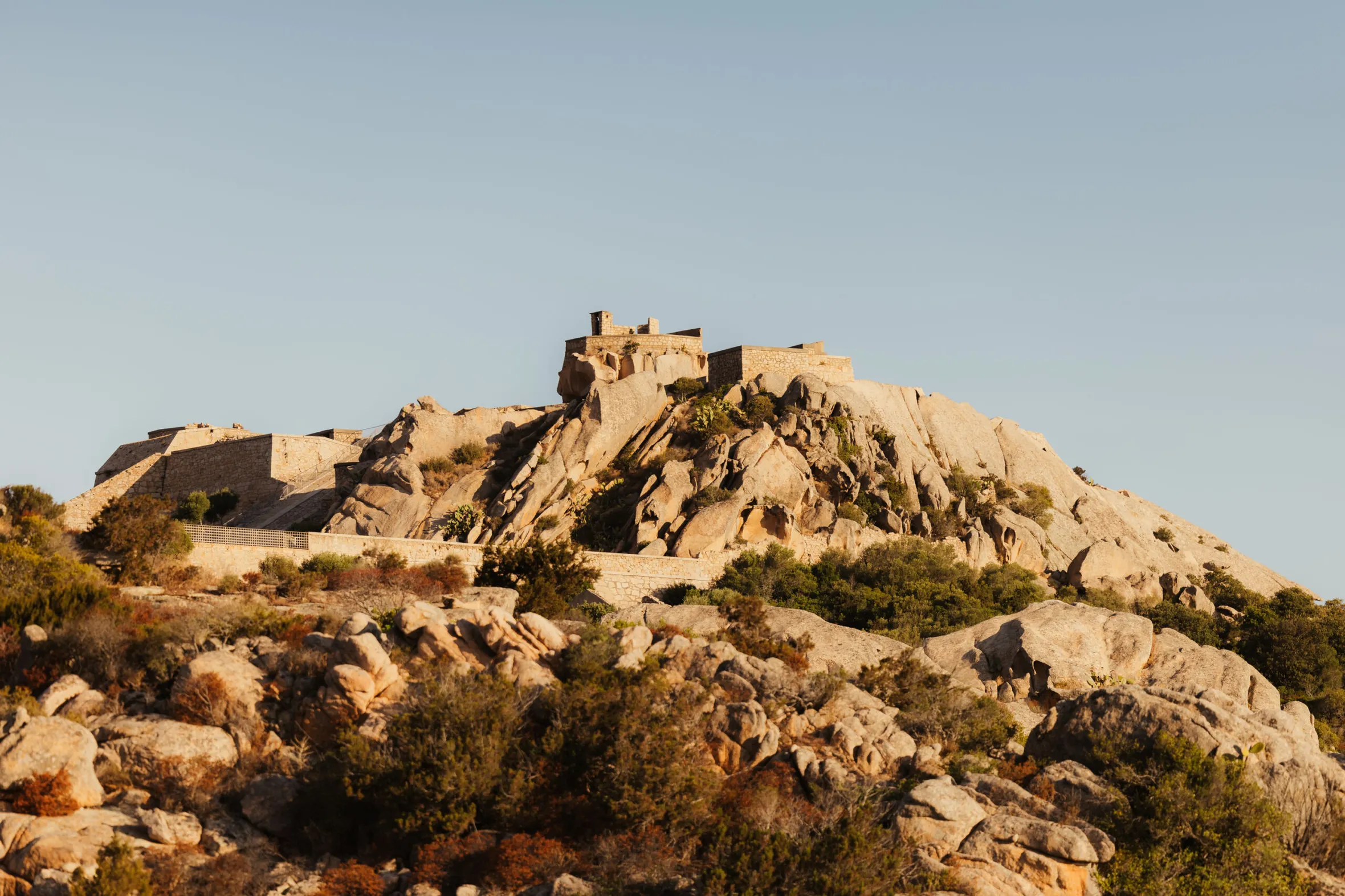 Ancient stone fortress atop a rocky hill, surrounded by sparse bushes, under a clear blue sky. Warm lighting gives the scene a historic, serene feel.