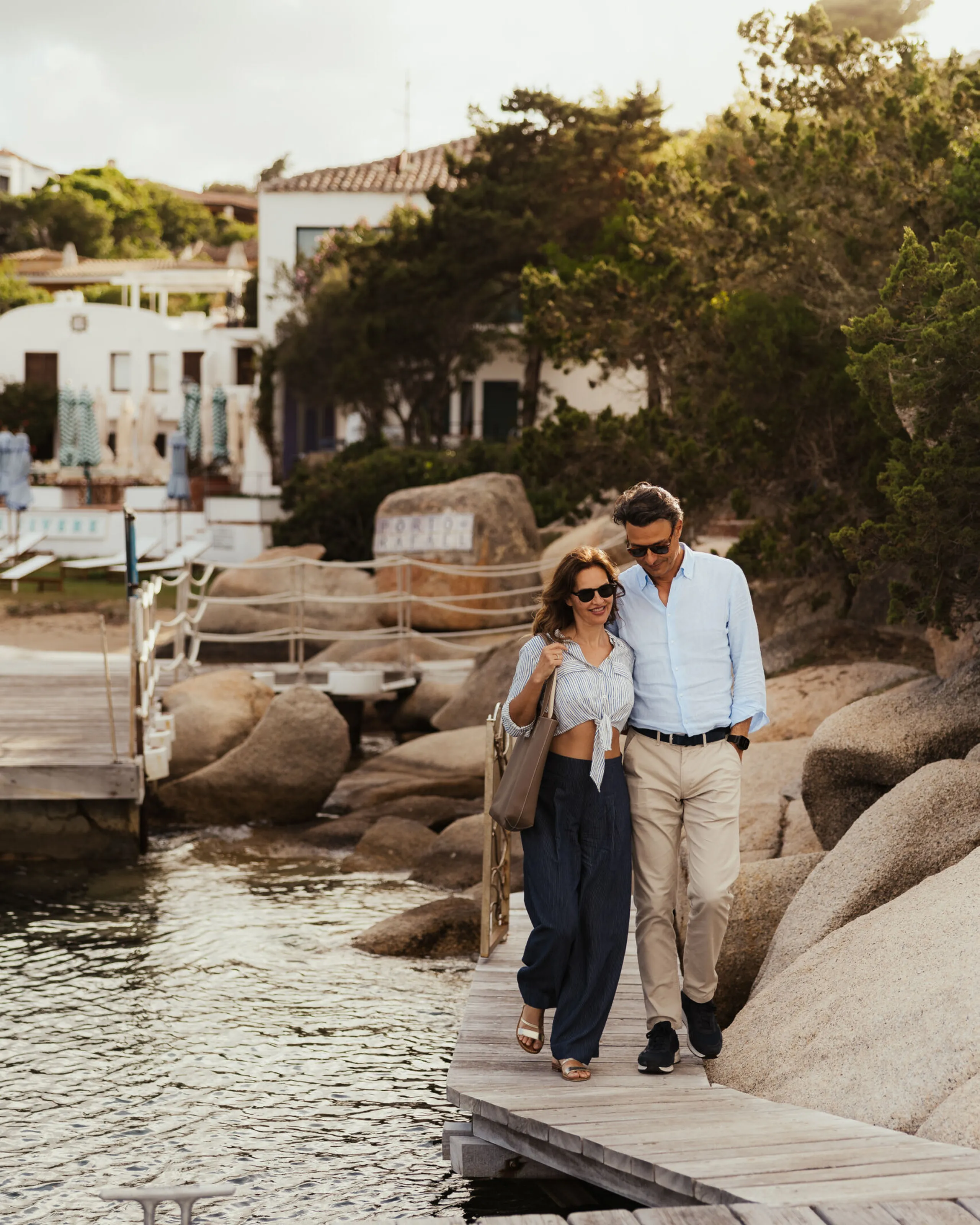 A man and woman stroll along a wooden dock beside calm waters, enjoying a sunny day at Porto Rafael.