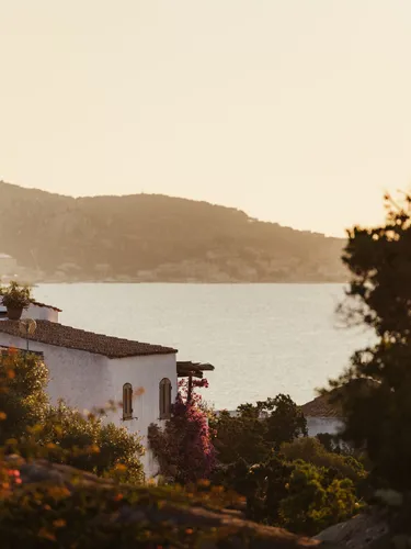 A serene coastal scene at sunset, featuring a white building with a tiled roof surrounded by lush greenery, and a calm sea with distant hills.
