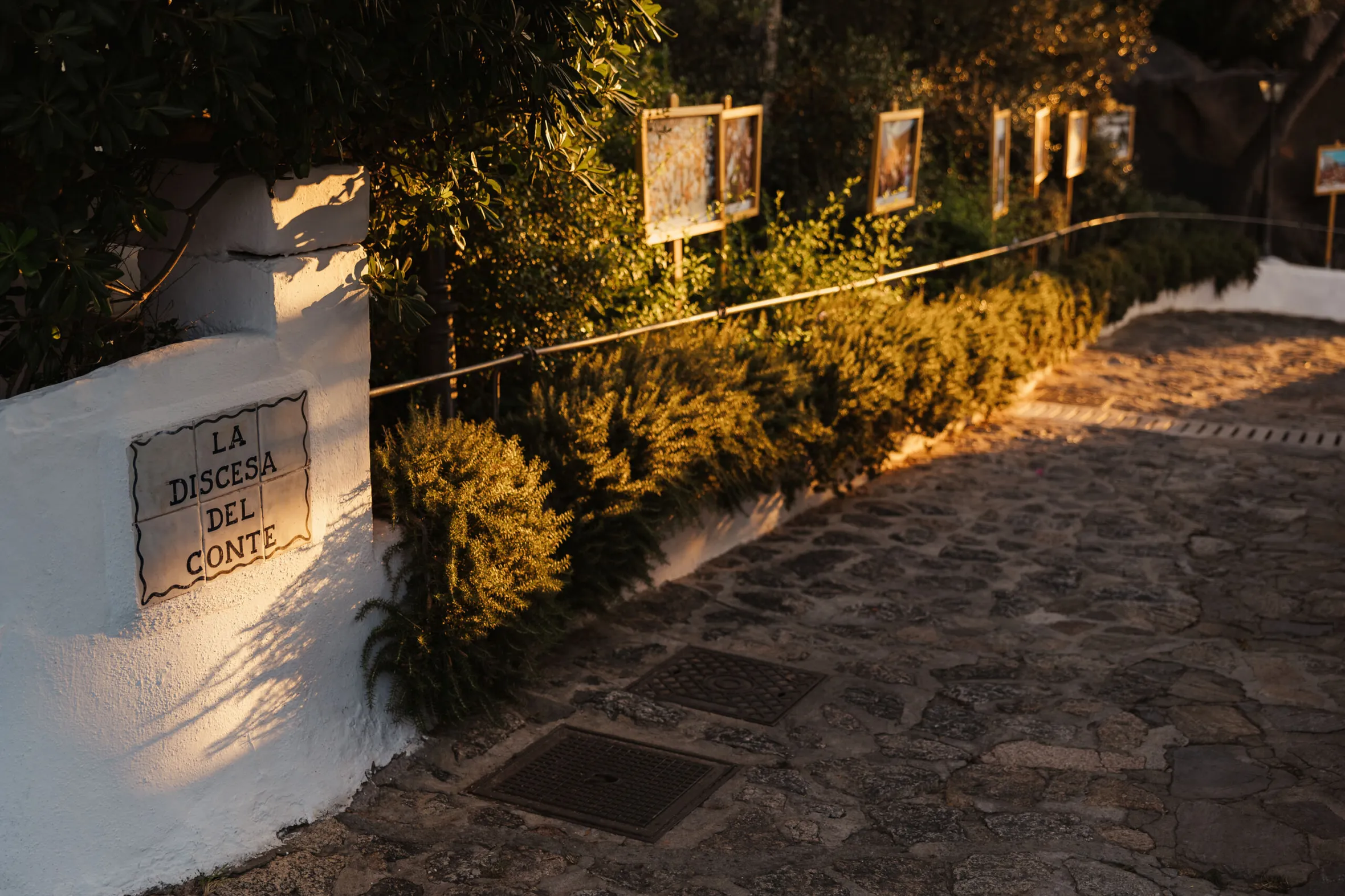 A sunlit stone pathway lined with greenery and framed paintings on stands. A white wall has a sign reading "La Discesa Del Conte," creating a serene ambiance.