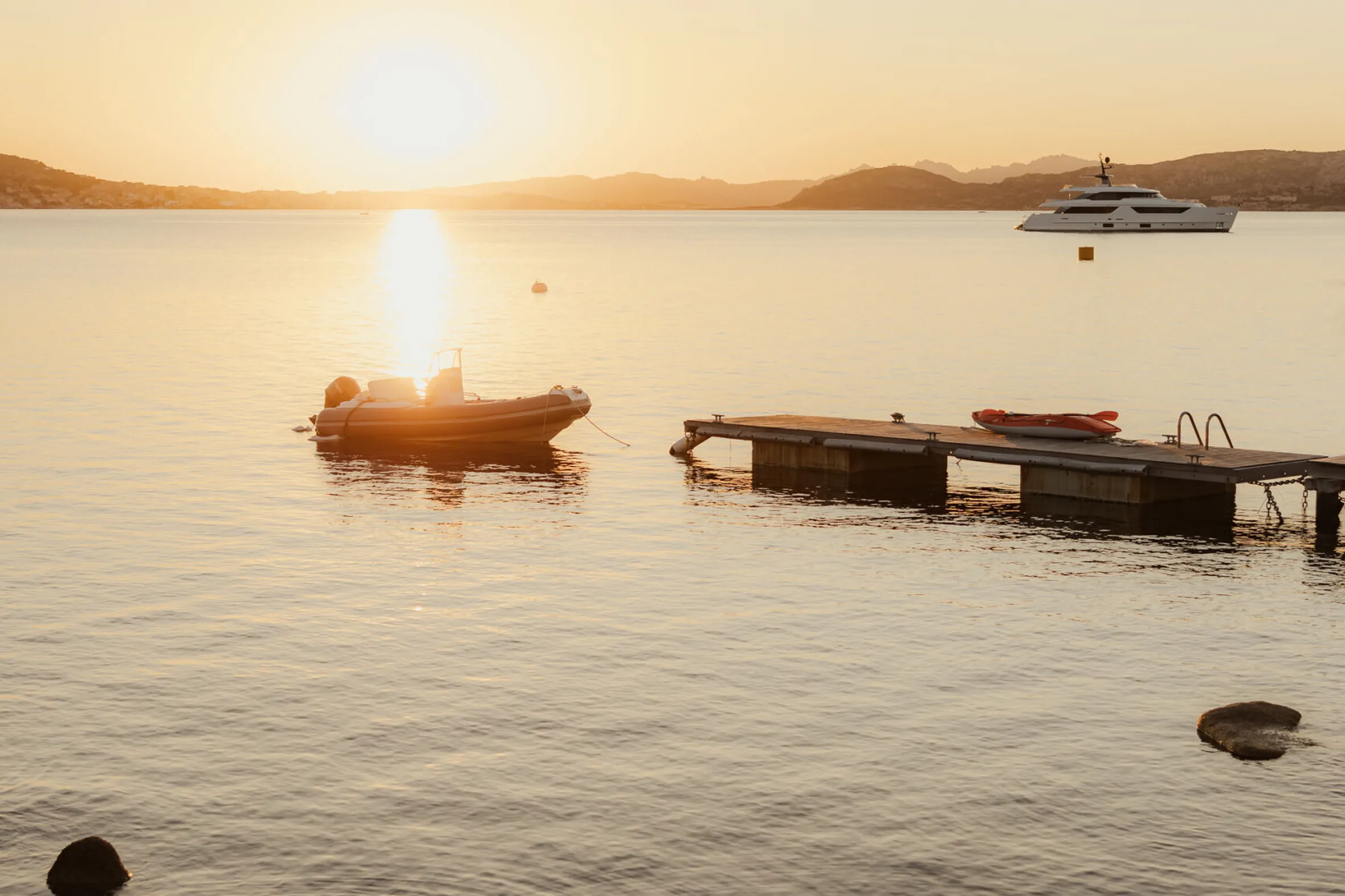 Serene sunset over calm water with a small boat near a wooden dock in the foreground. A yacht is visible in the distance. Tranquil ambiance.