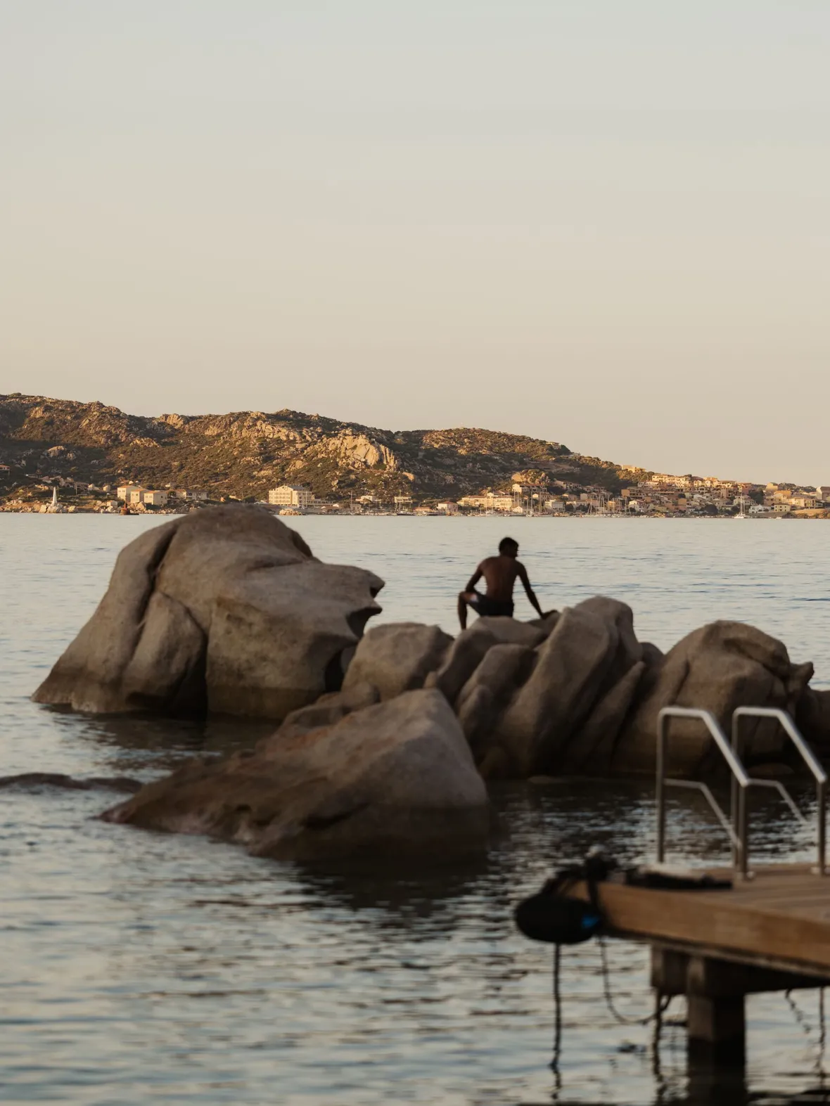A person sits on large coastal rocks with a distant cityscape and mountains under a clear sky at sunset. A wooden pier with a ladder is on the right.