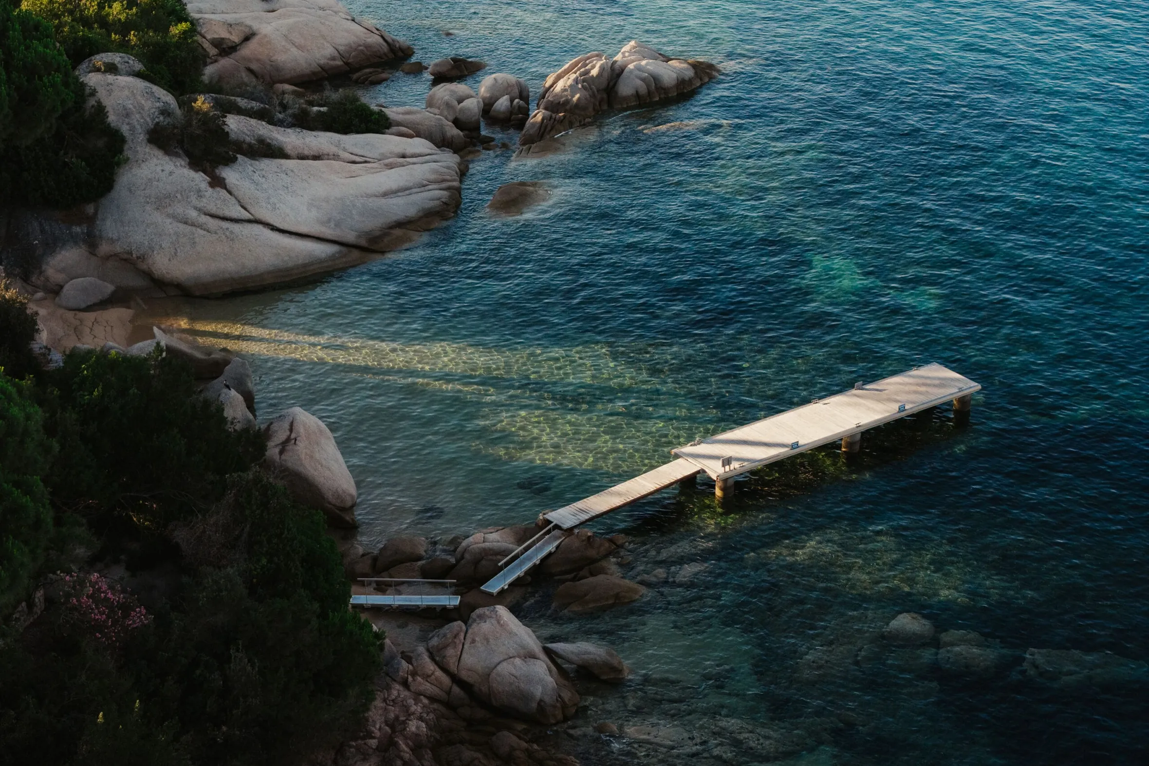 A wooden pier extends over clear, blue-green water, surrounded by large rocks and lush green bushes; the scene conveys tranquility and natural beauty.