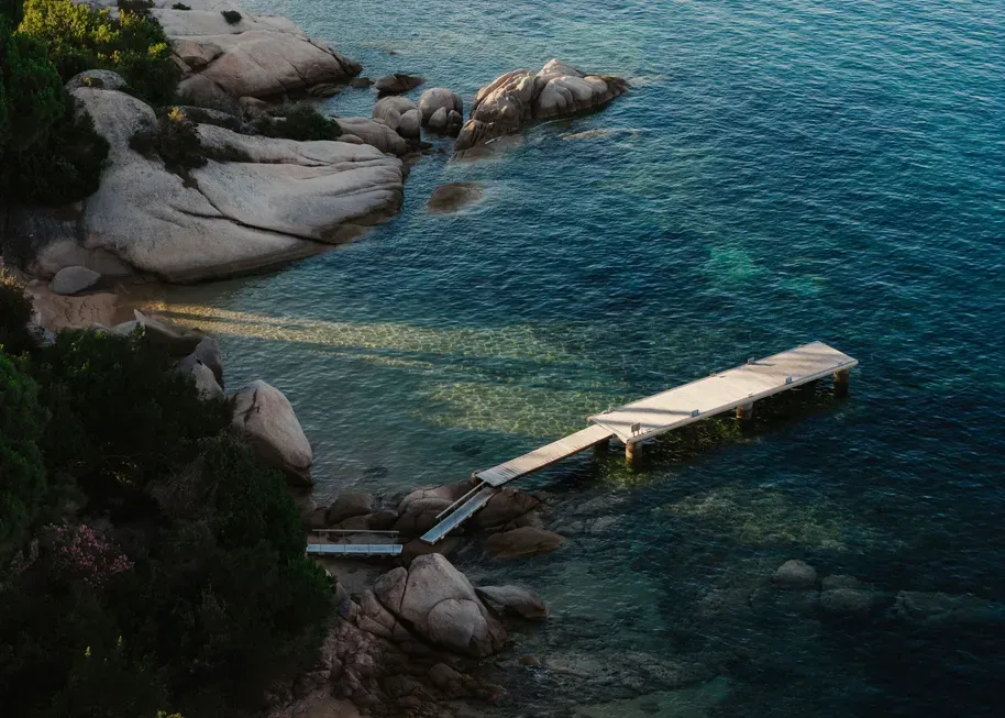 A wooden pier extends over clear, blue-green water, surrounded by large rocks and lush green bushes; the scene conveys tranquility and natural beauty.