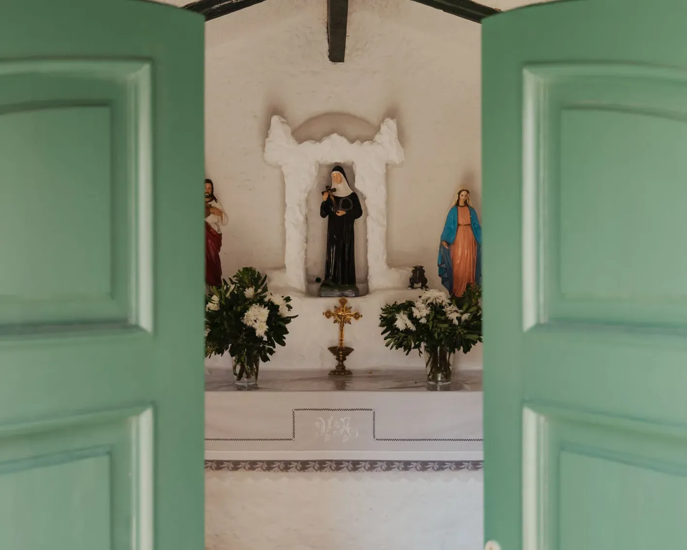 View through partially open green doors into a small chapel with religious statues, floral arrangements, and a tiled floor, conveying a serene atmosphere.