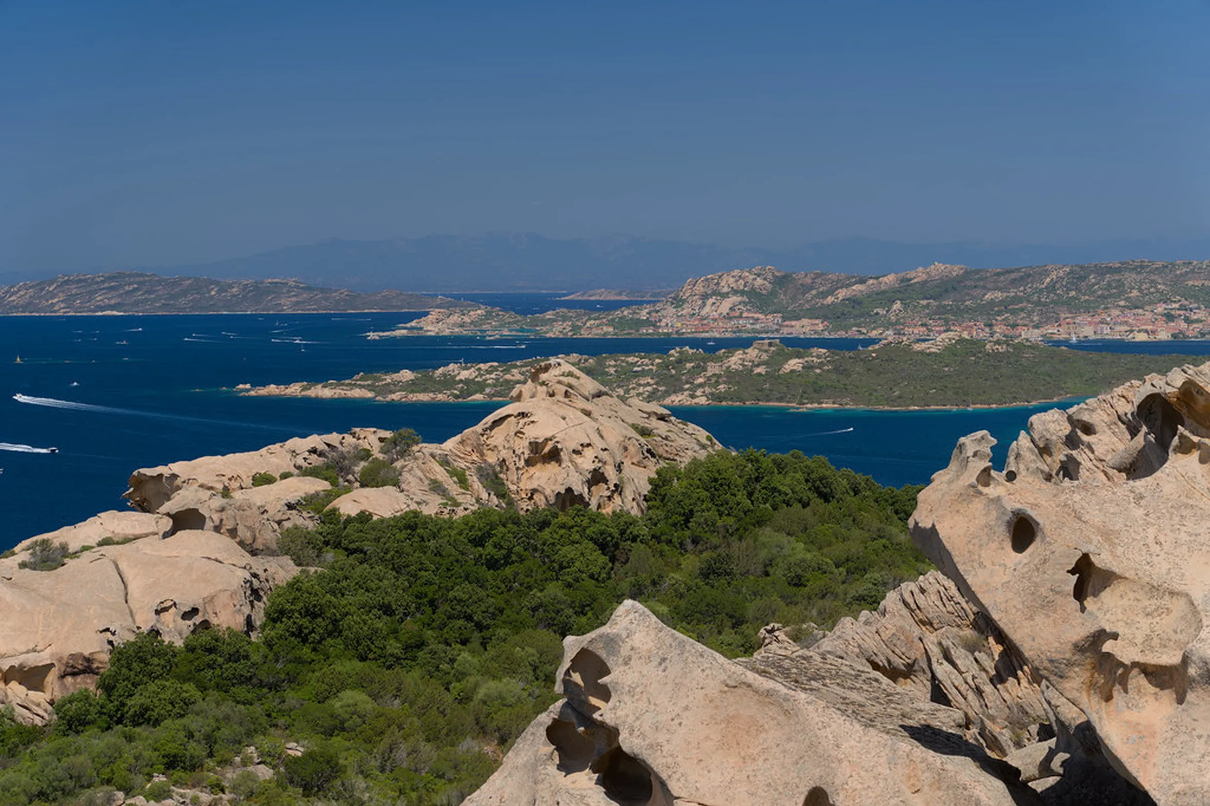 Scenic coastal view with rugged rocks, lush greenery, and a deep blue sea. Boats glide across the water under a clear, sunny sky. Tranquil and expansive.