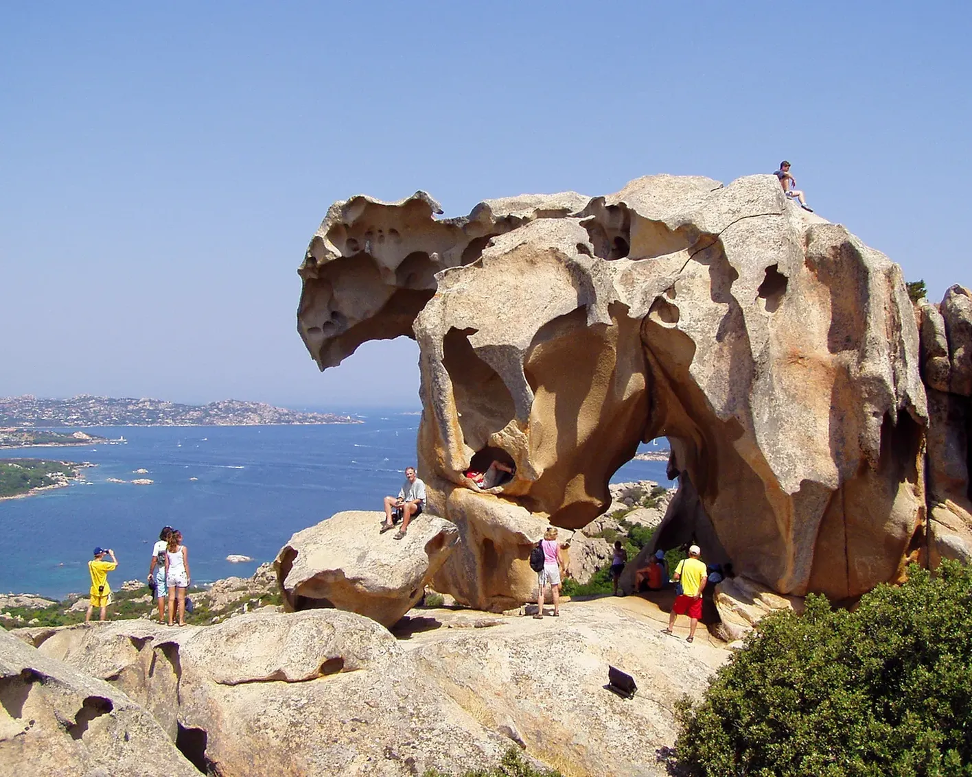 Spectacular rock formation on a sunny day with people exploring and relaxing on it. Background shows a panoramic view of the blue sea and coastline.
