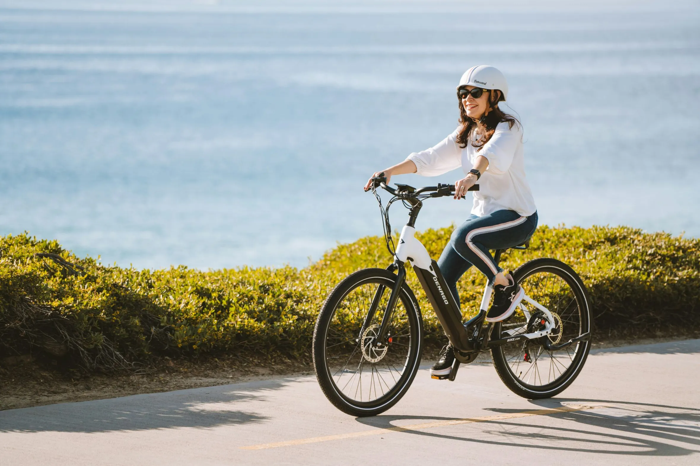 A woman in a helmet rides an electric bike along a coastal path. She appears happy and relaxed, with ocean waves and greenery in the background.