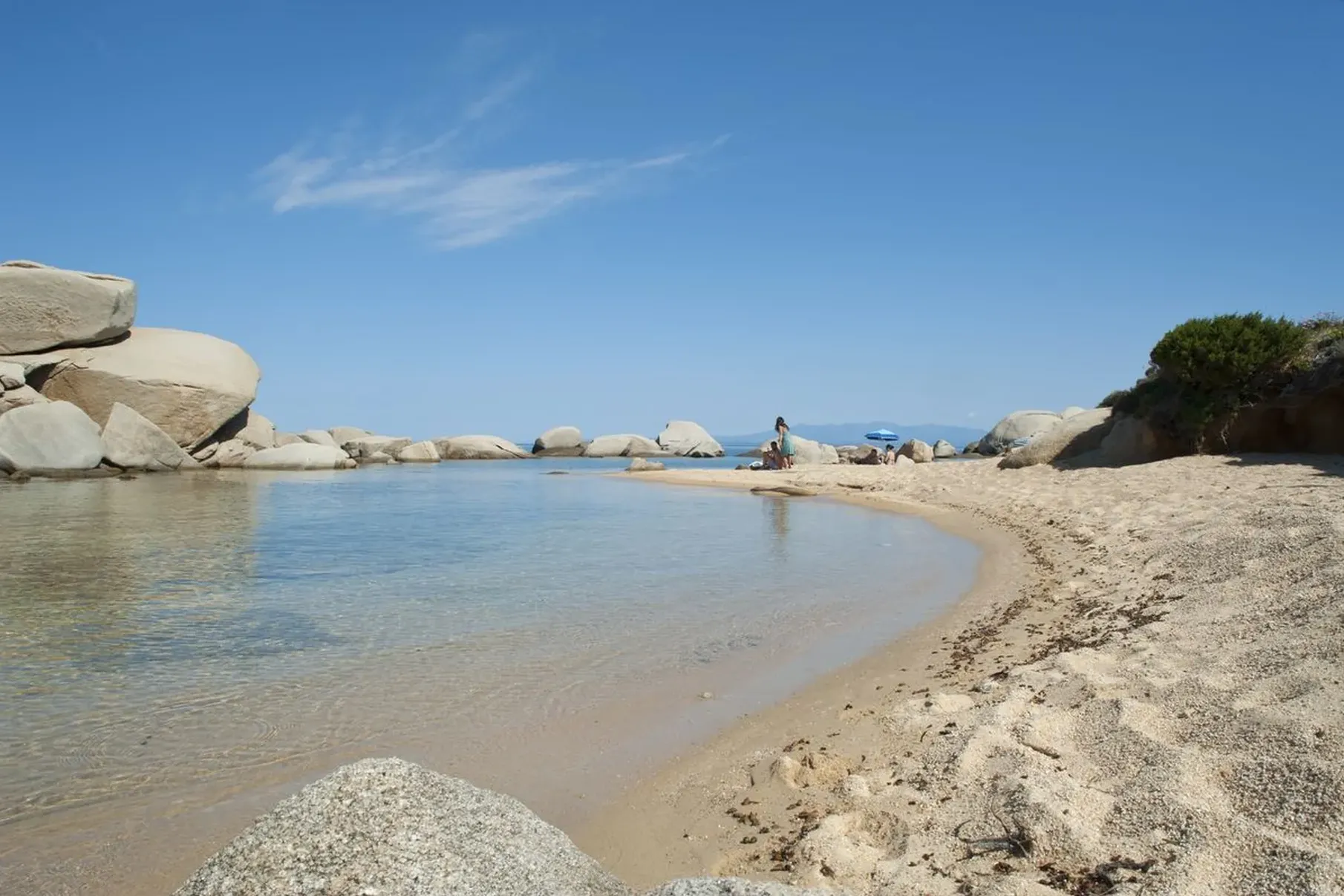 Sandy beach with clear, shallow water and large rocks under a bright blue sky. A person sits near the water's edge, evoking a peaceful, serene atmosphere.