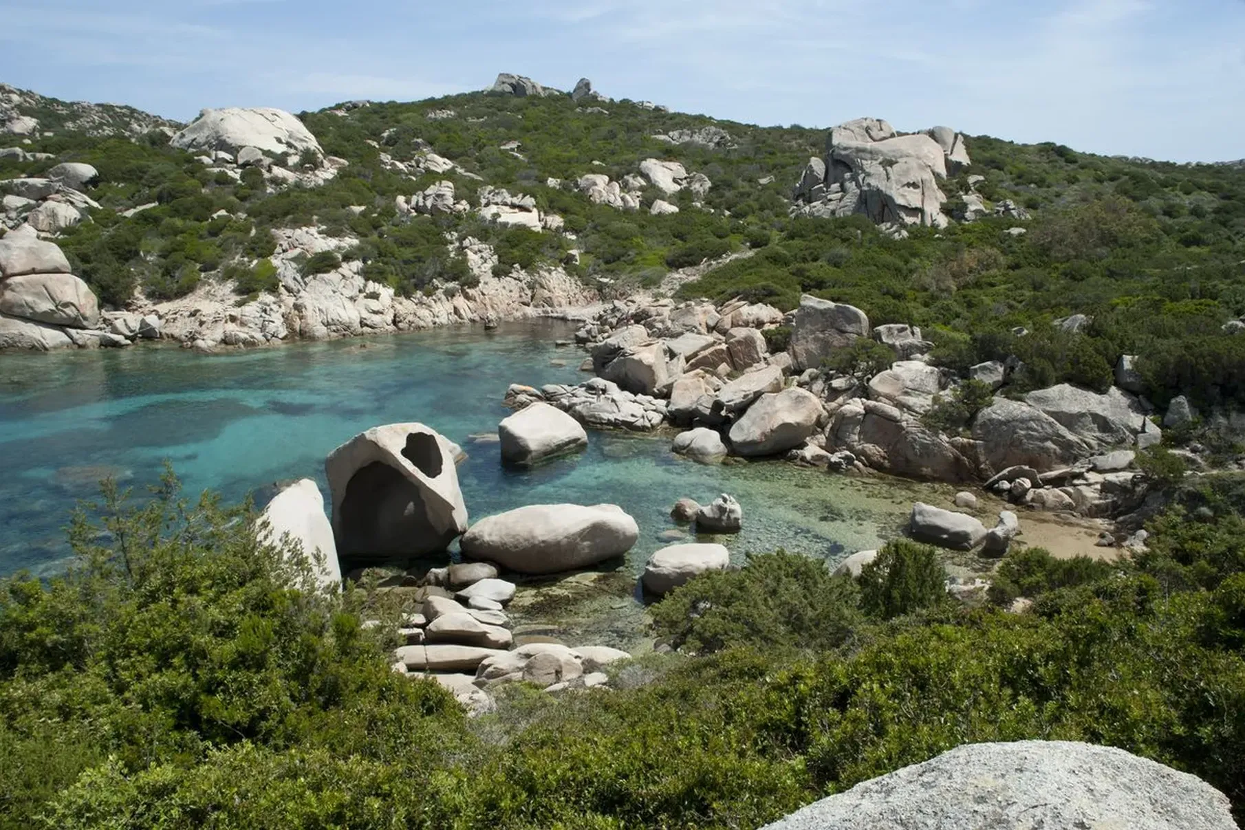 Rocky coastline with clear turquoise water, large boulders, and lush green vegetation under a bright blue sky, conveying a serene, natural beauty.