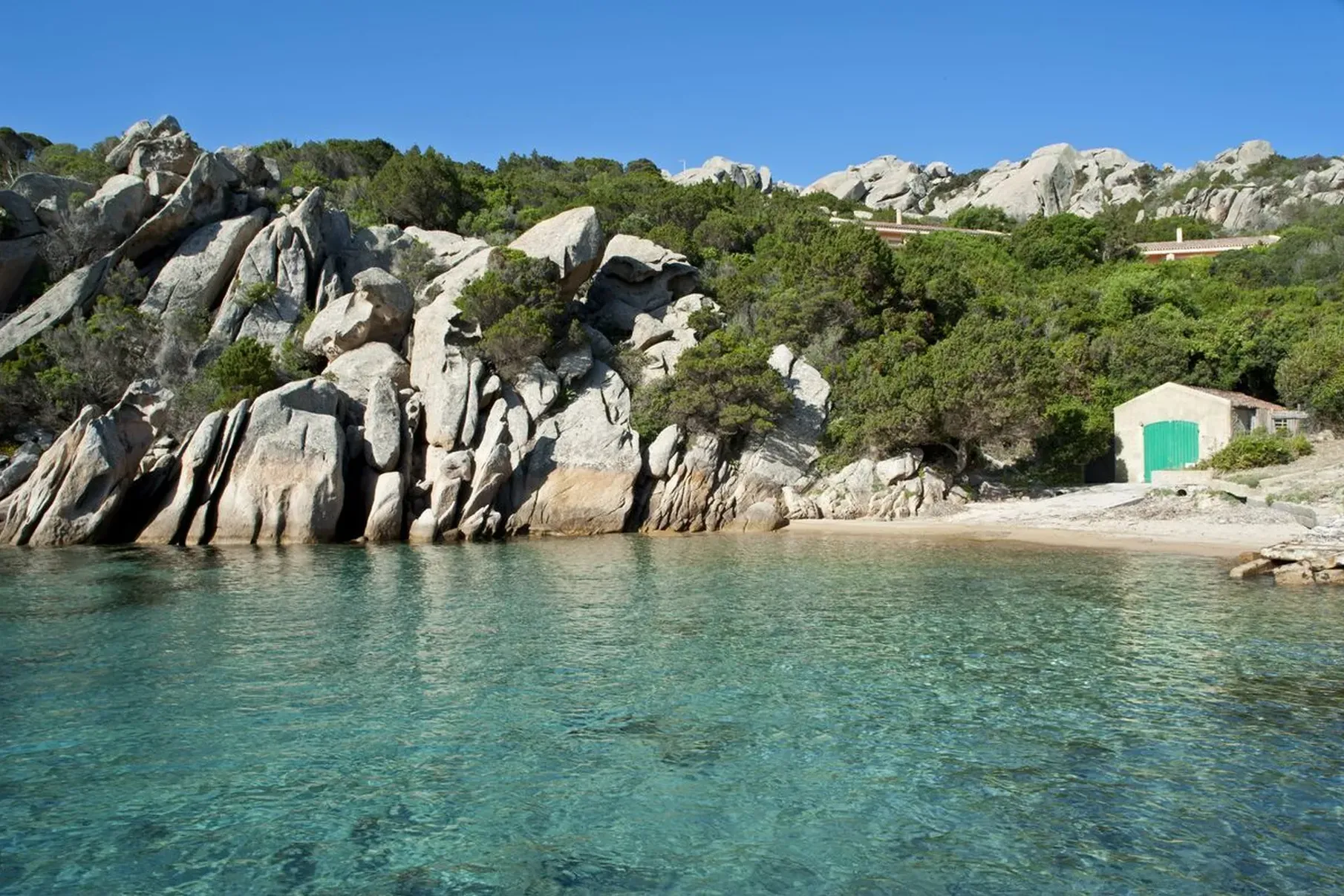 Rocky coastline with clear turquoise water and a small sandy beach. A rustic stone building with a green door sits among lush greenery under a clear blue sky.