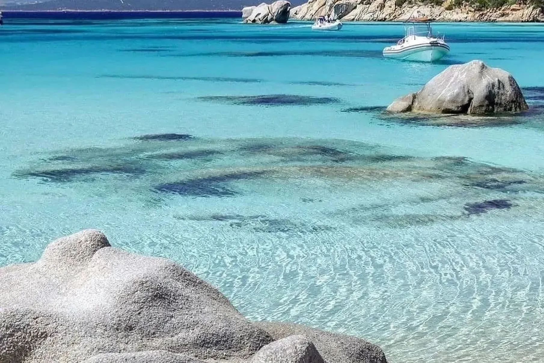 Clear turquoise waters stretch across the tranquil beach with smooth rocks in the foreground. A boat floats in the distance under a bright blue sky.