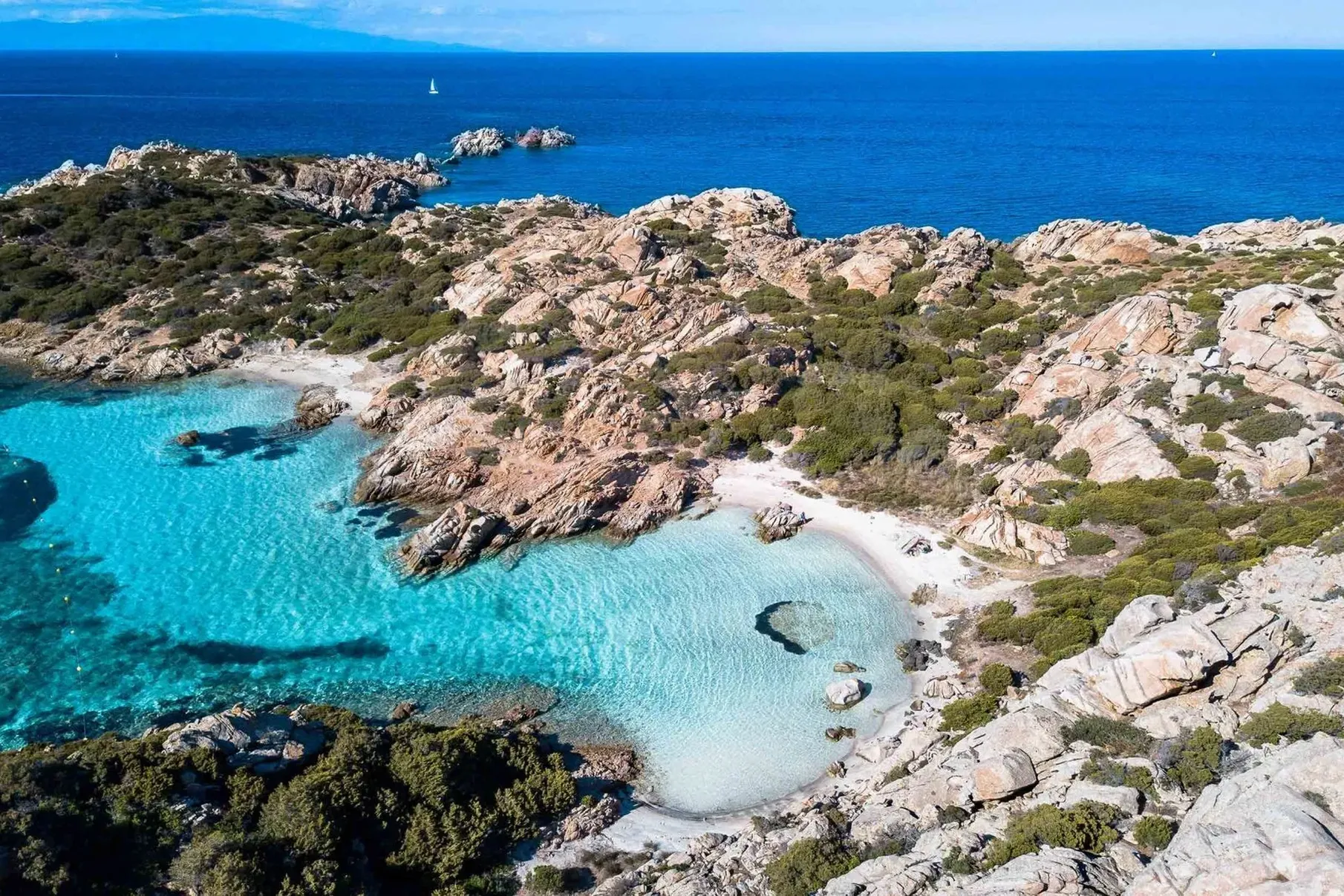 Aerial view of a serene coastal landscape with clear turquoise waters and sandy coves surrounded by rocky terrain and lush green shrubs under a bright blue sky.
