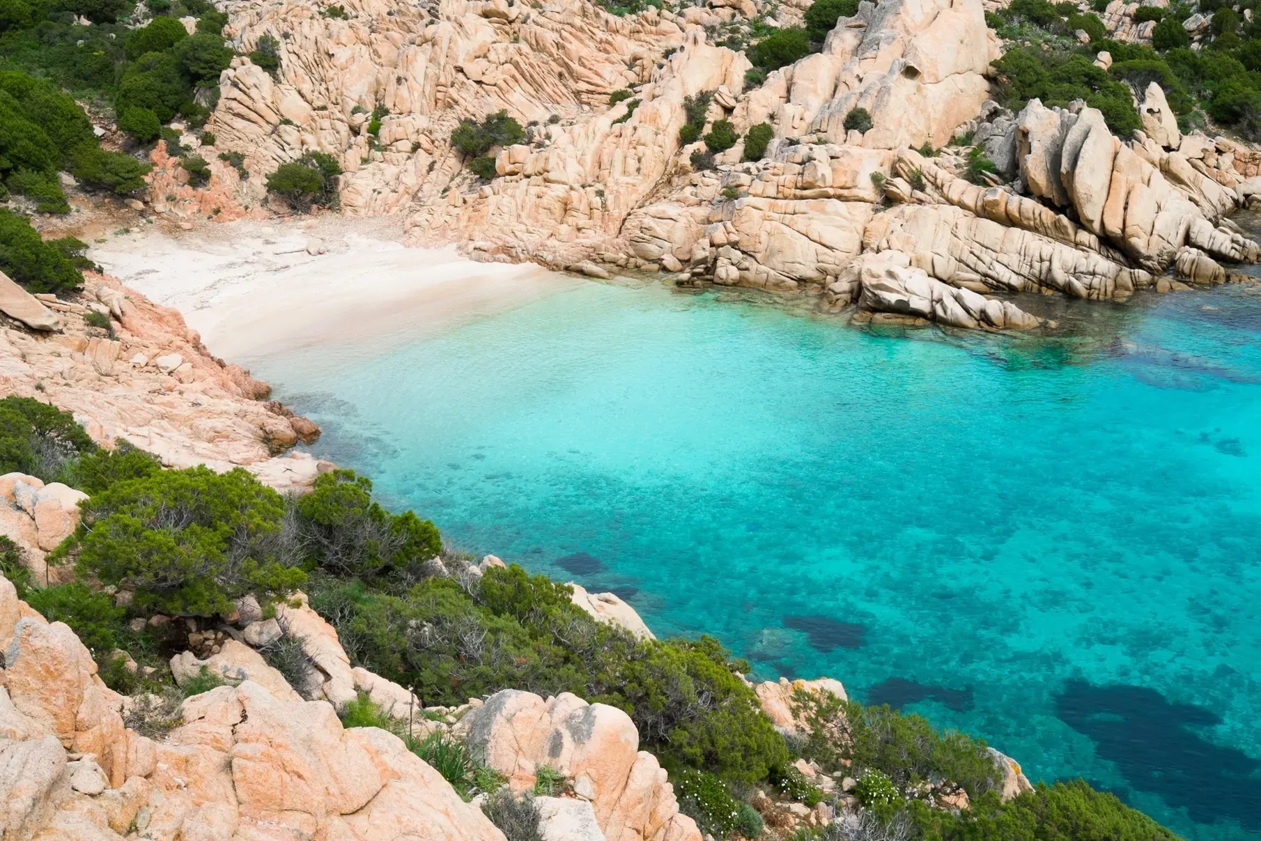 Rocky coastal landscape with clear turquoise water and a small sandy beach. Surrounding green shrubs add a vibrant contrast to the serene scene.