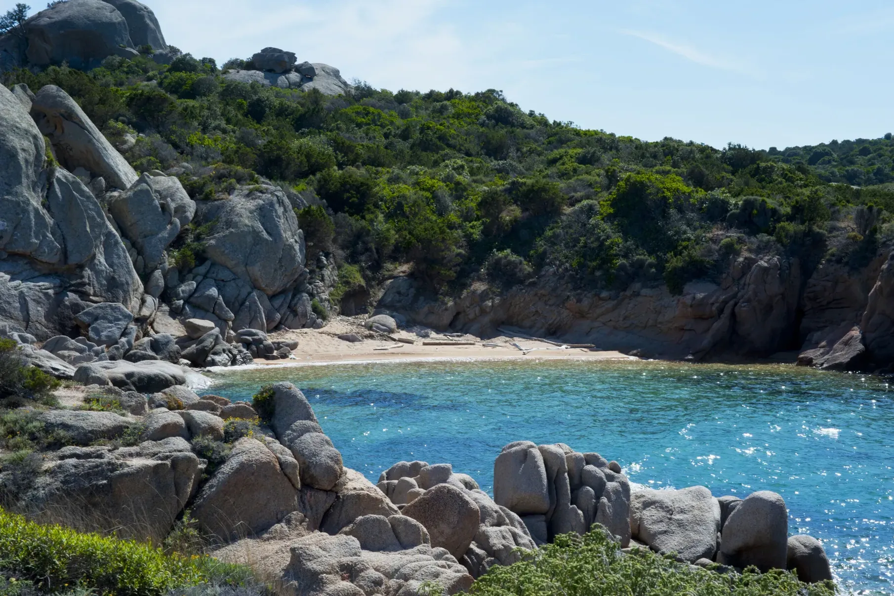 Rocky cove with vibrant turquoise water, surrounded by lush green hills and large boulders. A serene, secluded beach under a clear blue sky.
