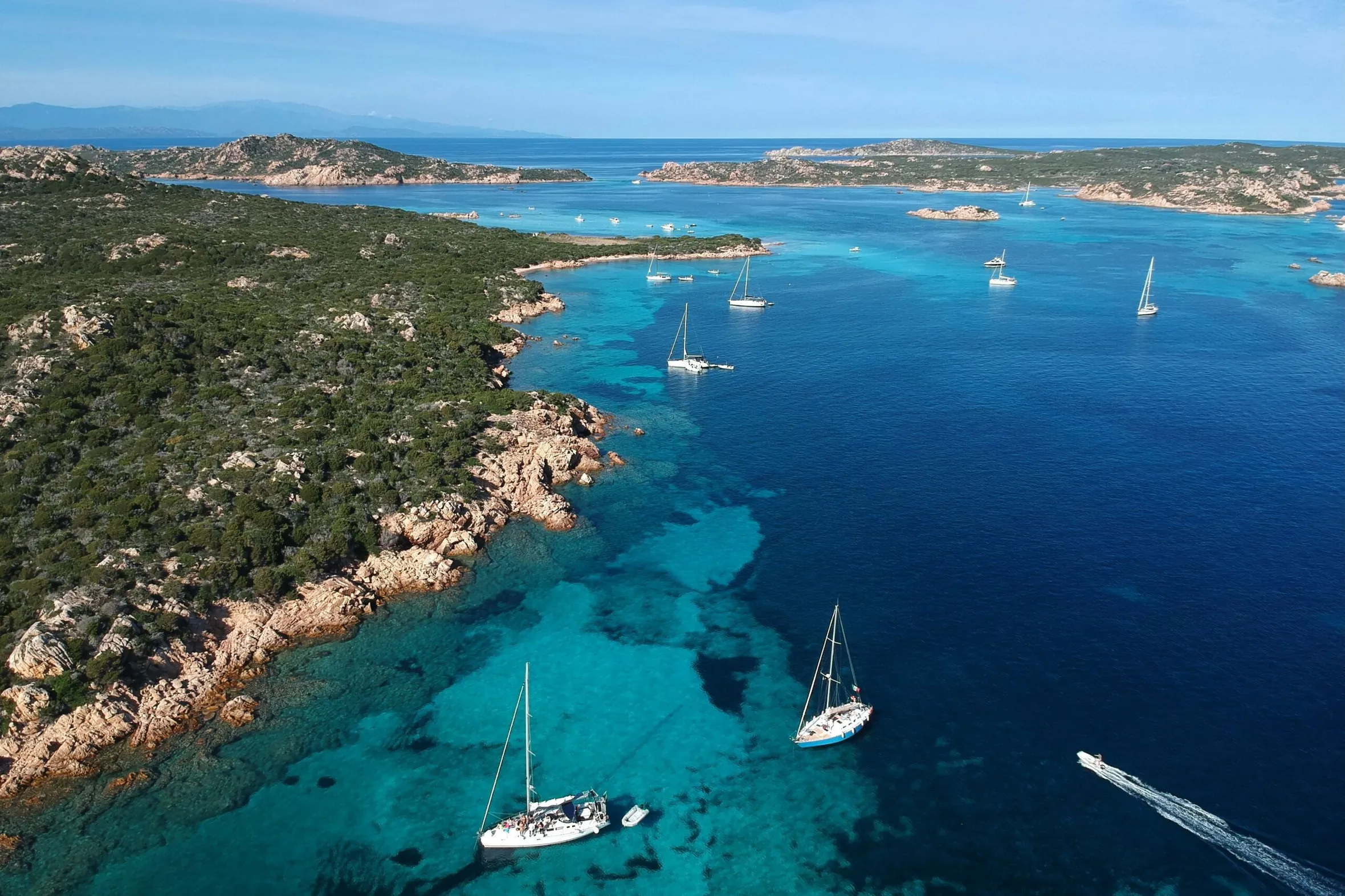 Vue aérienne d’un paysage côtier avec des eaux turquoise et des voiliers. Le littoral rocheux est recouvert d’une végétation luxuriante. La scène est calme et pittoresque.