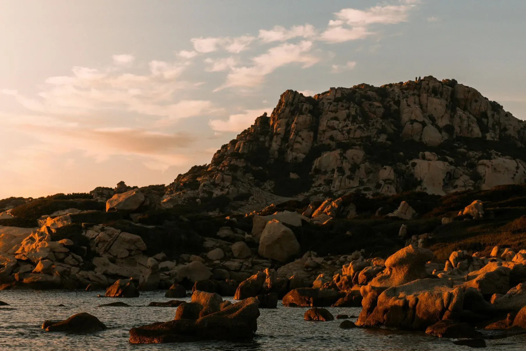 Côte rocheuse au coucher du soleil avec de grandes falaises escarpées sous un ciel parsemé de nuages, reflétant de chaudes nuances dorées sur une eau calme, créant une scène paisible.