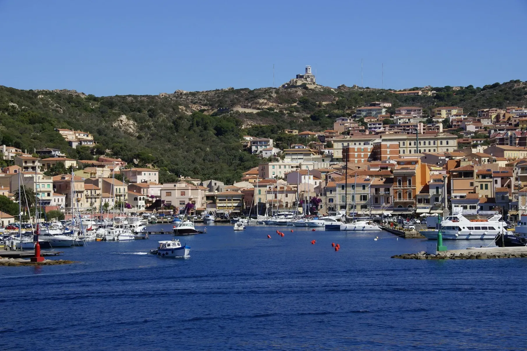 Ville côtière aux bâtiments colorés et aux collines luxuriantes. Des bateaux sont amarrés dans le port bleu sous un ciel dégagé, créant une atmosphère à la fois sereine et animée.